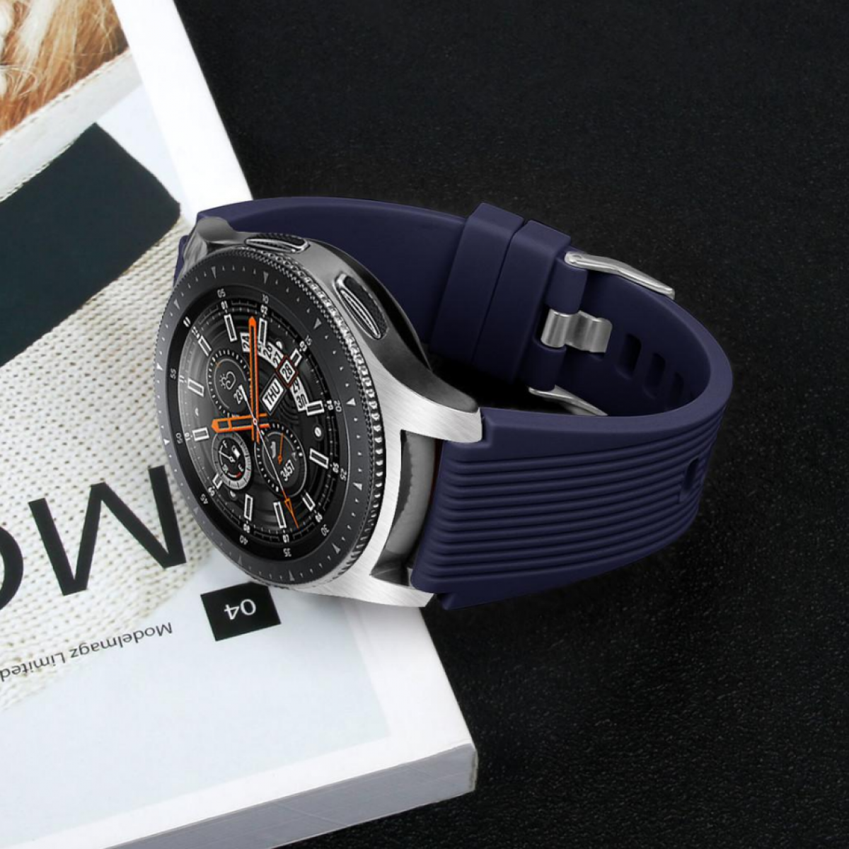 Eine Smartwatch mit dunkelblauem Armband, auf einem Magazin, dunkler Hintergrund.