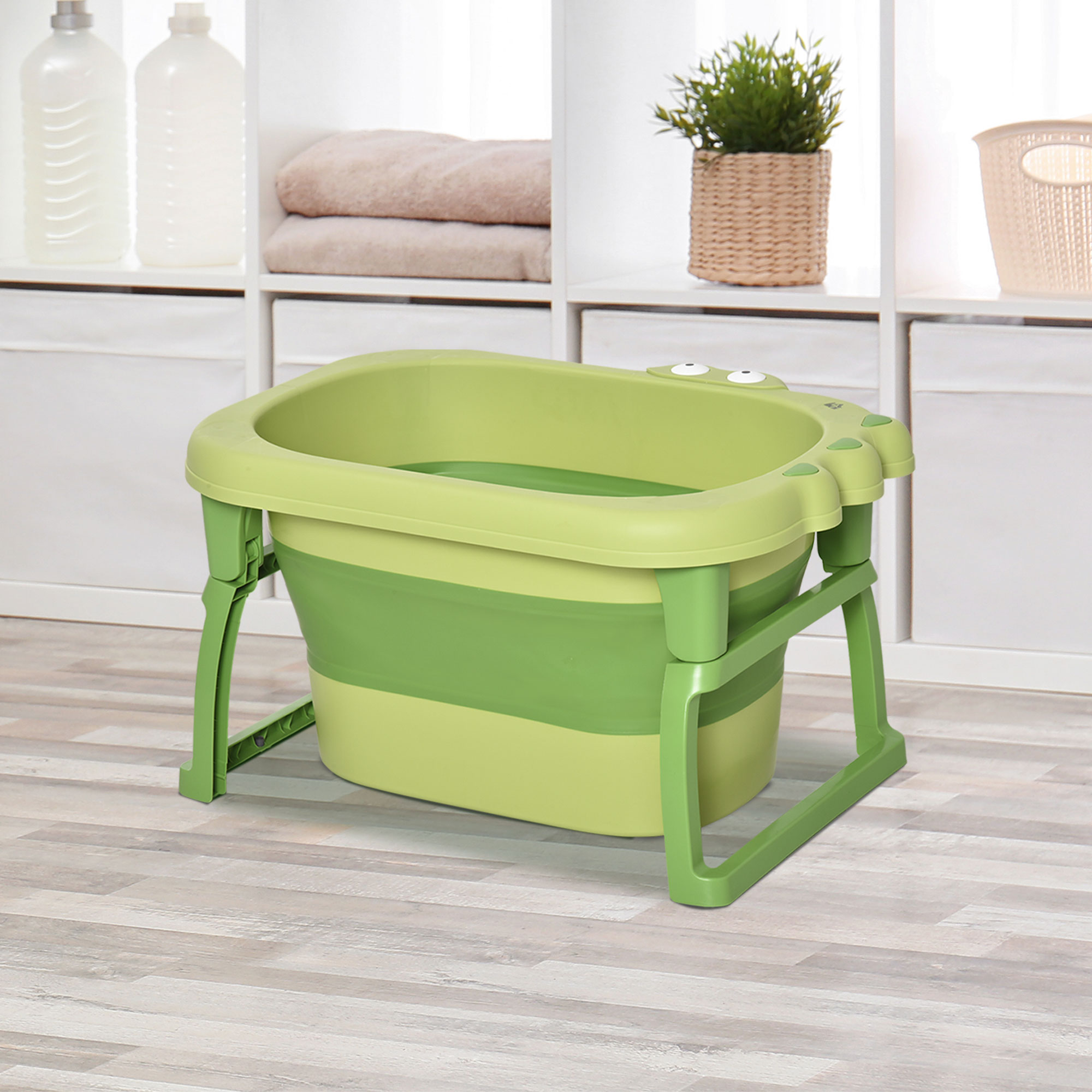 Una bañera plegable para bebés verde y verde claro con ojos de rana en un suelo de madera clara.