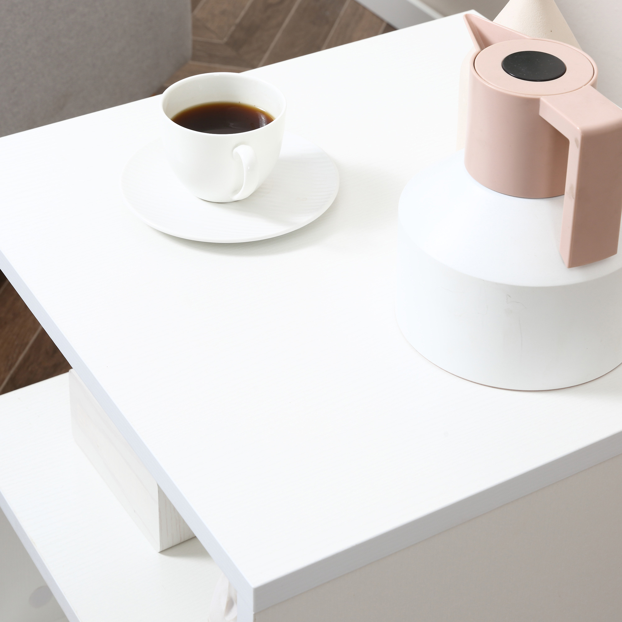 Una taza de café blanca está sobre un platillo blanco junto a una tetera rosa.