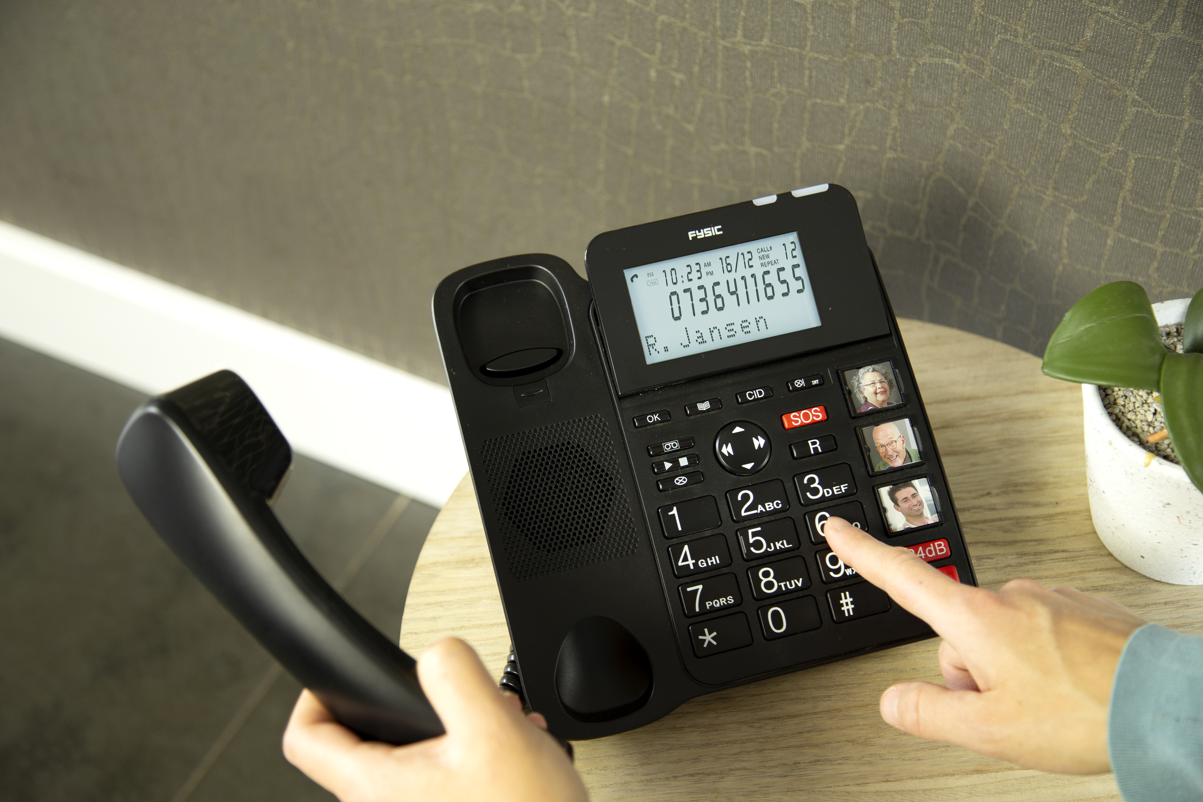 Téléphone noir avec grands boutons et écran affichant contacts et heure. Main tenant le combiné.