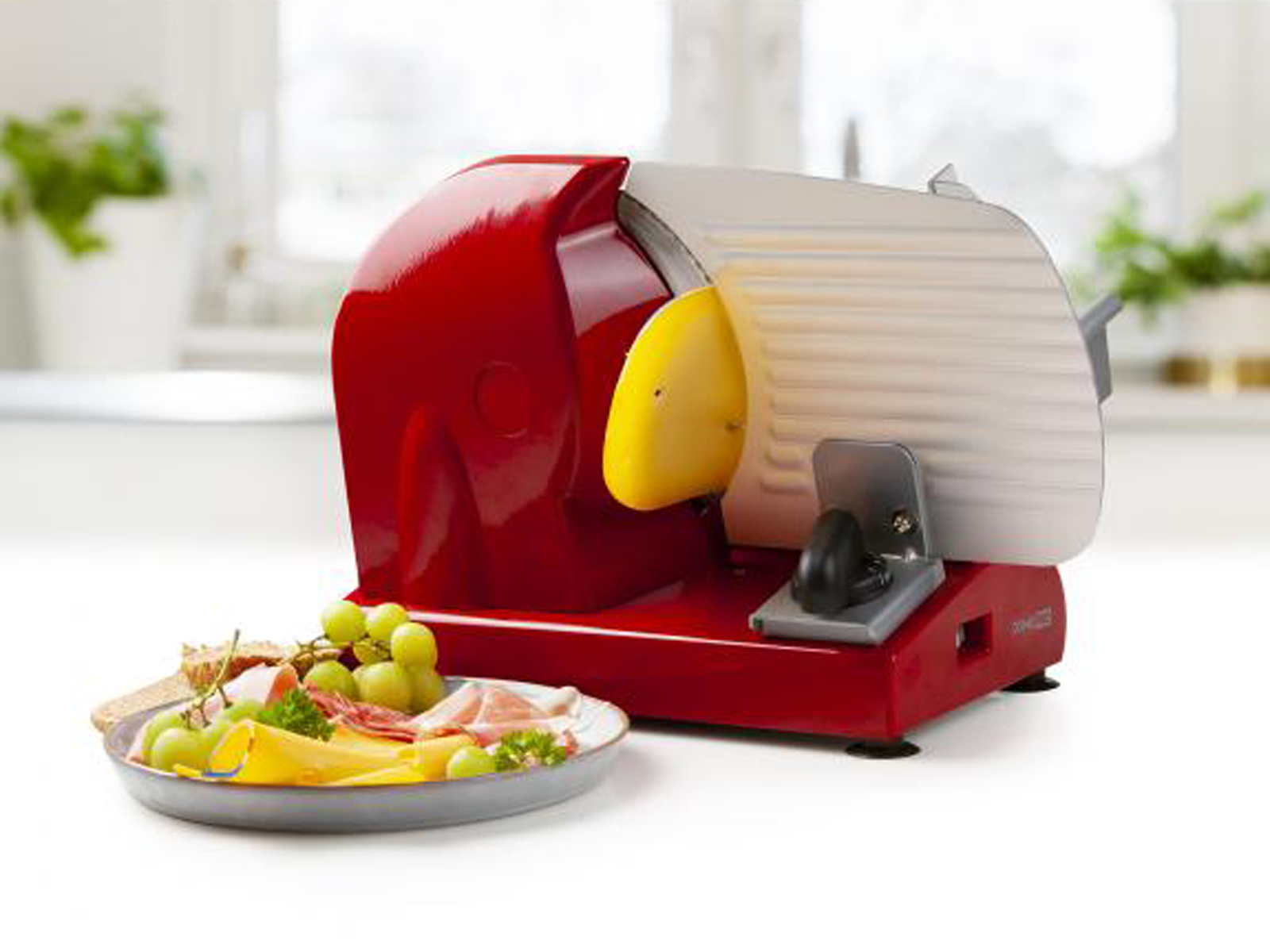 Ein roter Lebensmittelschneider mit gelber Führung, mit Käse, Trauben und Fleisch auf einem Teller.