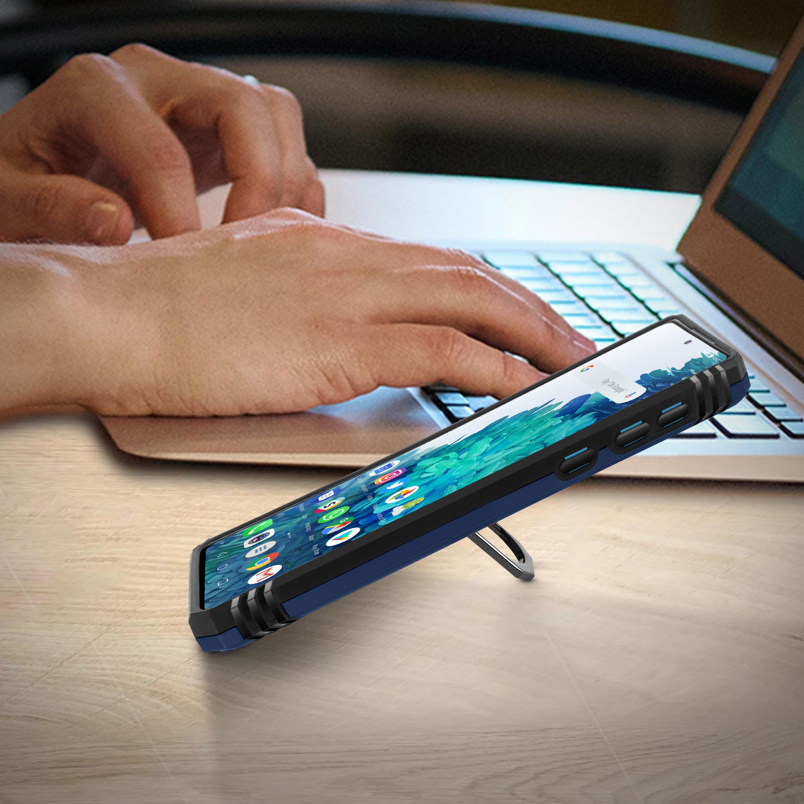 Persoon die een laptop gebruikt met een telefoonhoesje op een tafel. Telefoon staat met de standaard open.