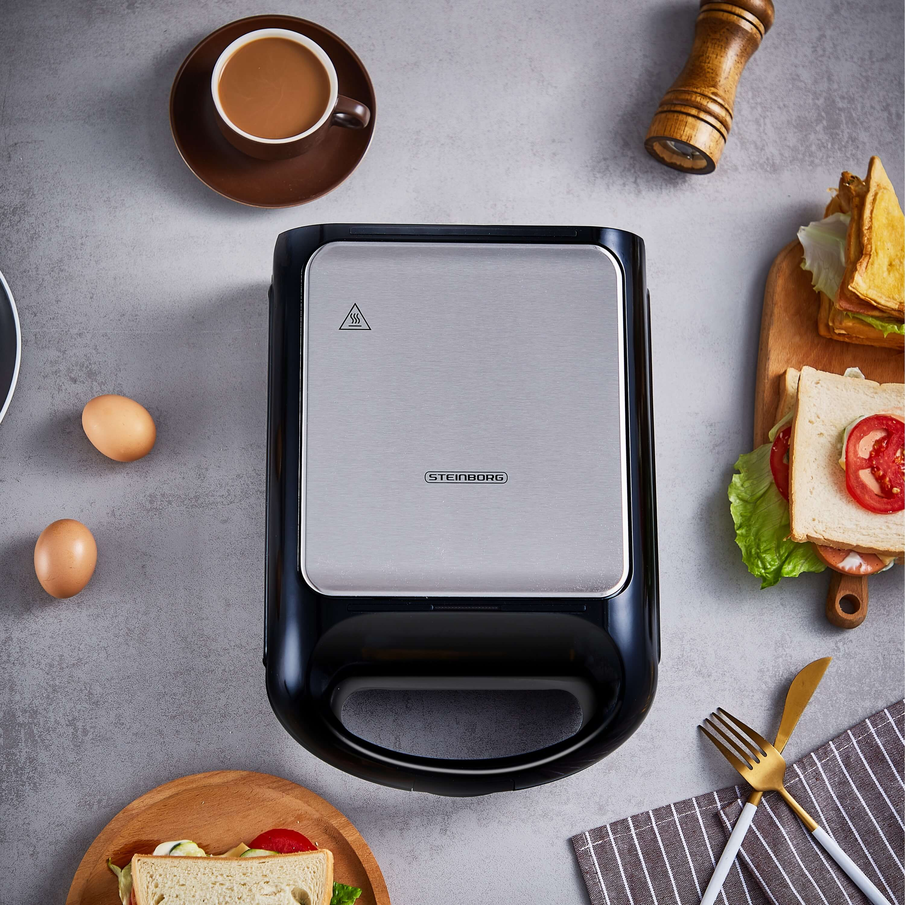 Ein Sandwichmaker mit schwarzer und silberner Oberseite. Zwei Eier, Kaffee, Sandwich und ein Holzbrett sind sichtbar.
