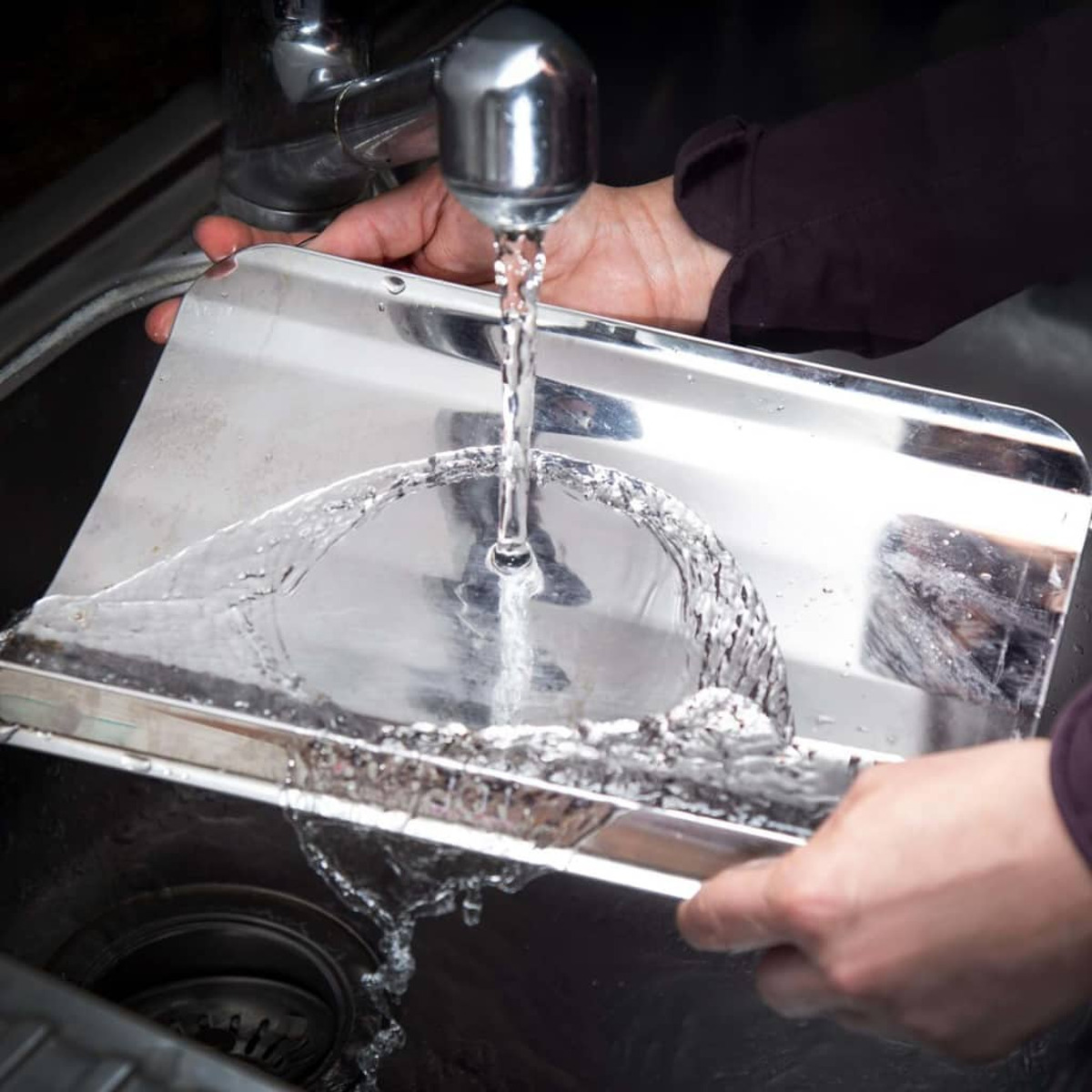 Mani che lavano un vassoio in acciaio inox sotto l'acqua corrente in un lavandino scuro.