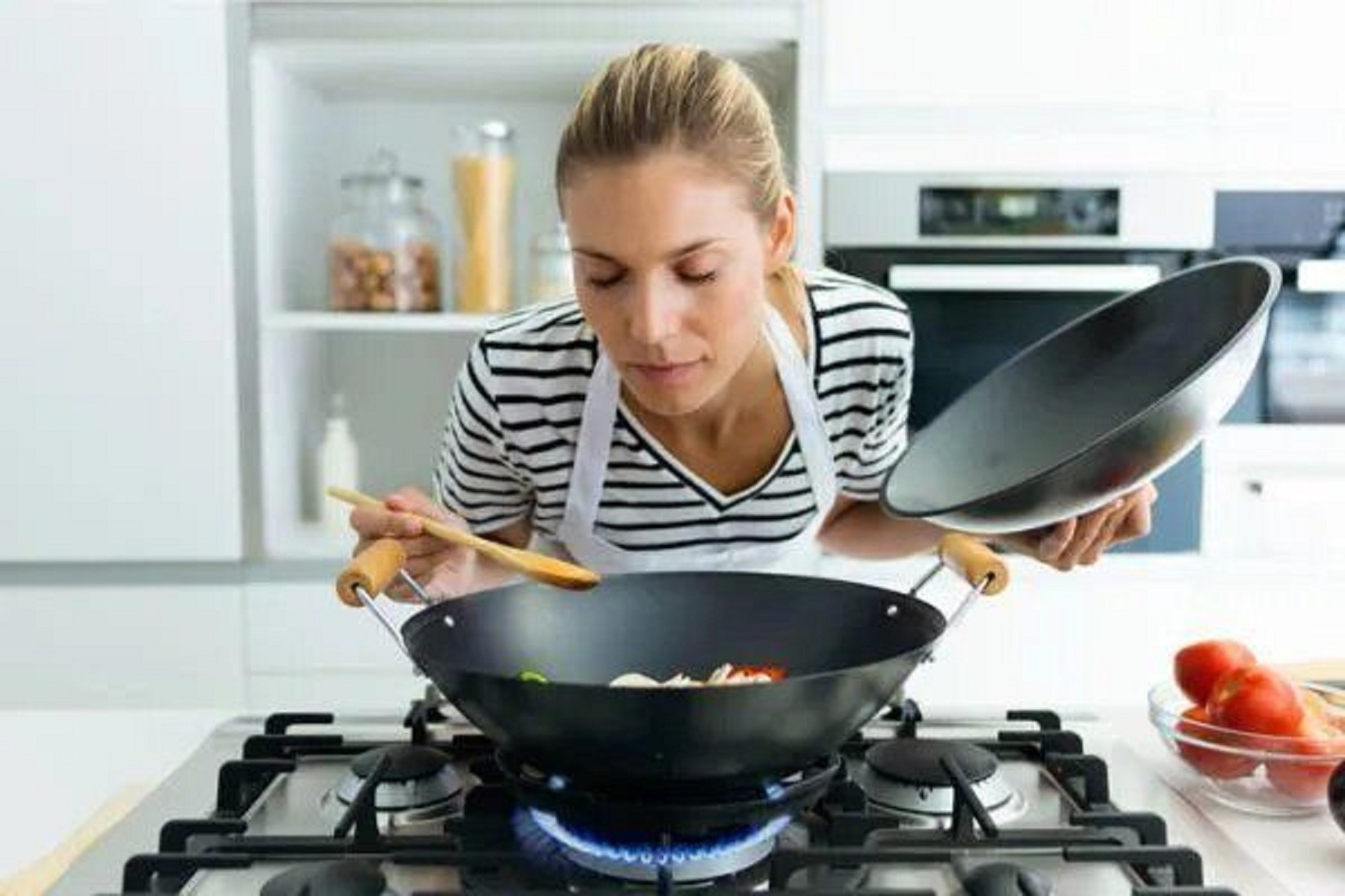Mujer en una cocina oliendo el contenido de un wok. Sostiene una cuchara de madera y una tapa.