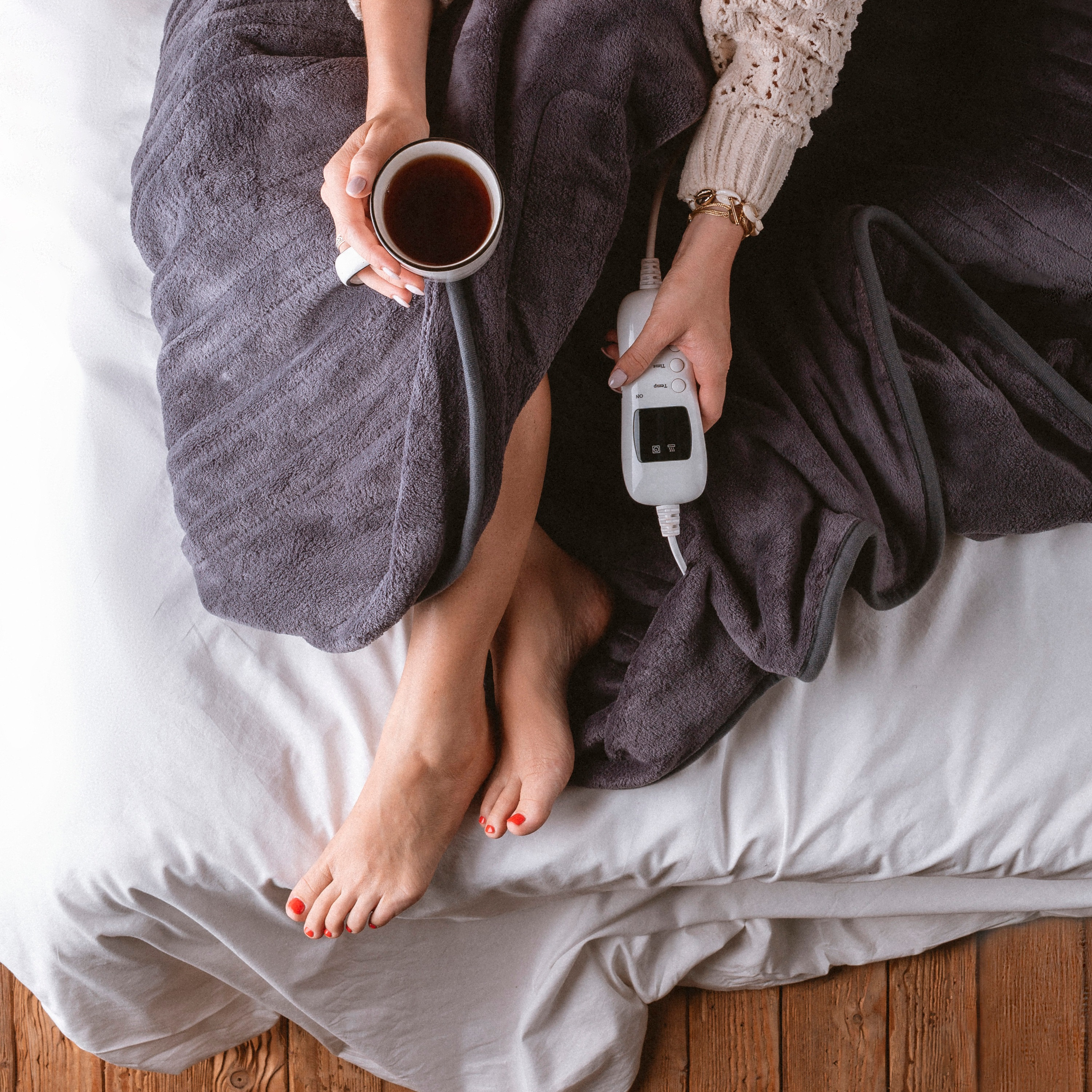 Donna con smalto rosso, avvolta in una coperta grigia, che tiene in mano una tazza di caffè e un telecomando.
