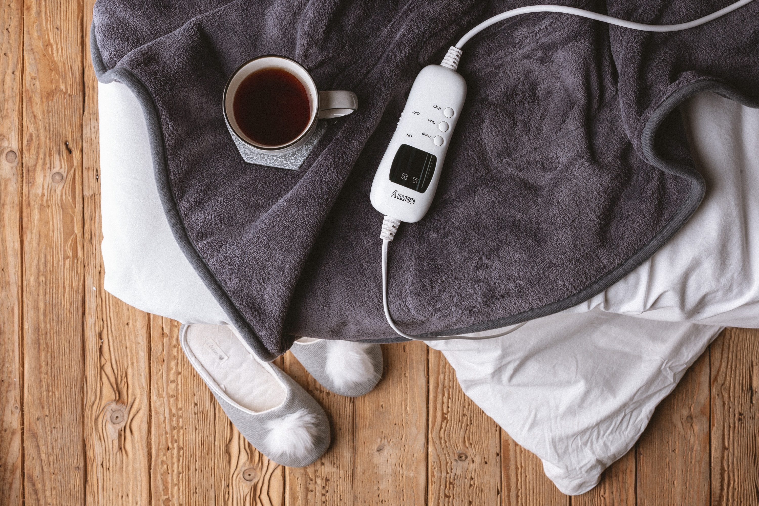 Vista dall'alto: coperta, tazza di tè, pantofole e controllo della coperta elettrica su legno.