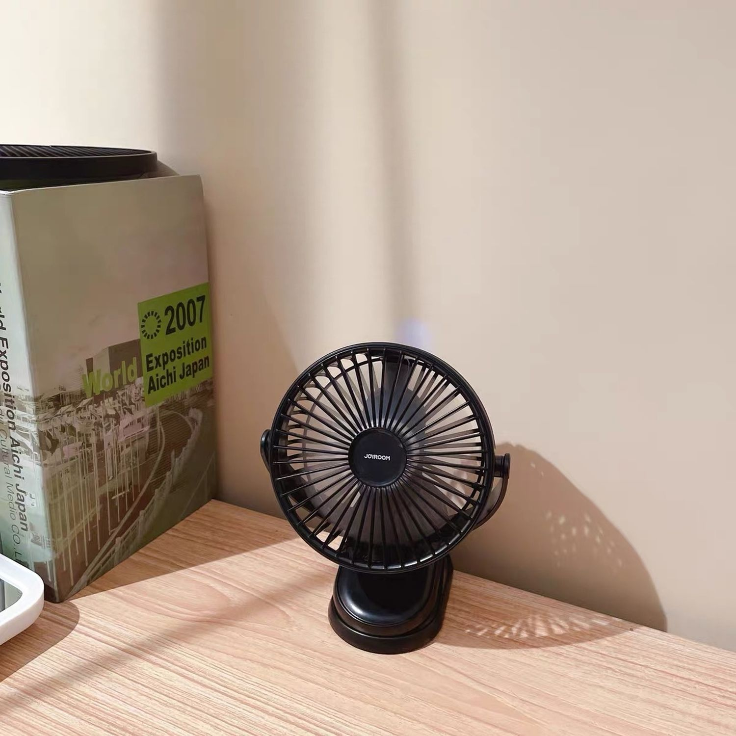 Schwarzer Tischventilator auf einem Holztisch. Links ist ein Buch und hinten die Wand.