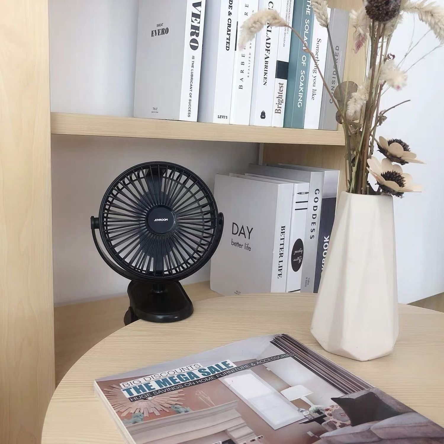 Ein kleiner Ventilator auf einem Tisch neben Büchern und einer Vase mit Blumen.