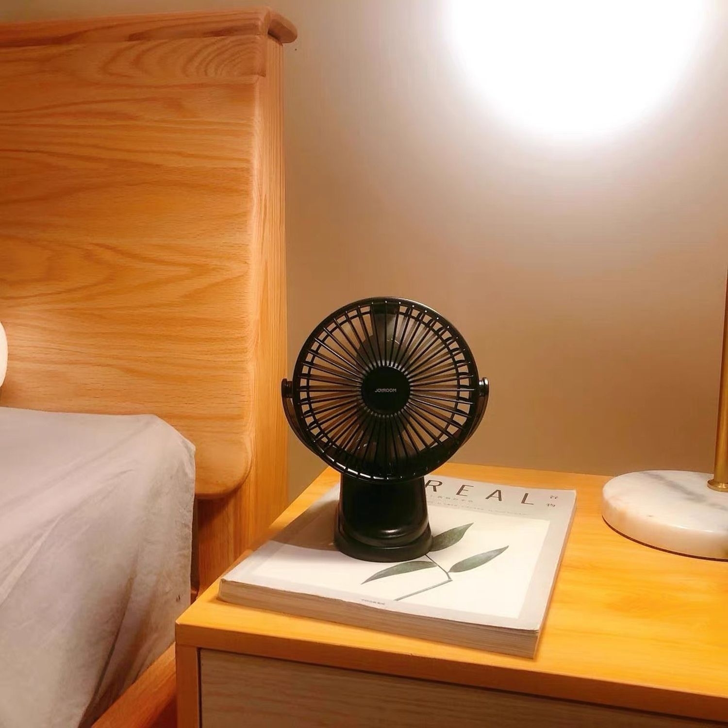 Ein kleiner schwarzer Ventilator auf einem Tisch mit einer Zeitschrift und einer Lampe. Holzbett im Hintergrund.
