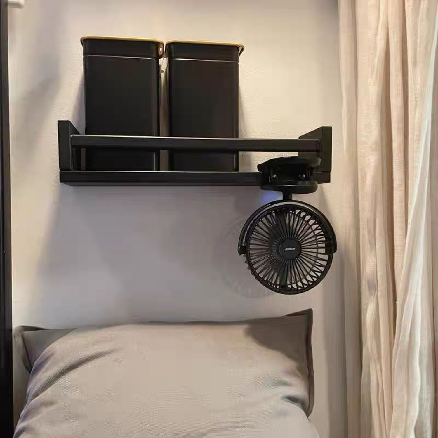 Ein an der Wand befestigtes Regal mit zwei schwarzen Behältern, einem Ventilator und einem Kissen auf dem Bett.