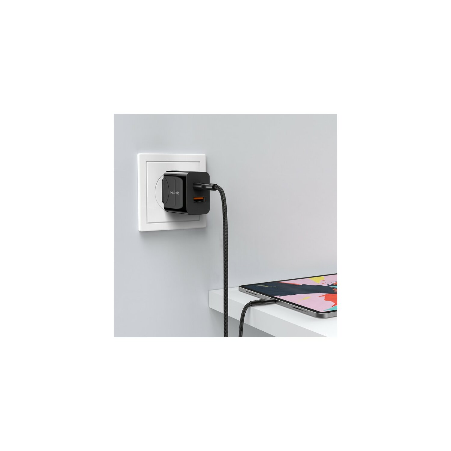 Ein schwarzes Ladegerät in einer Steckdose, mit einem Ladekabel, das mit einem Tablet auf einem weißen Regal verbunden ist.