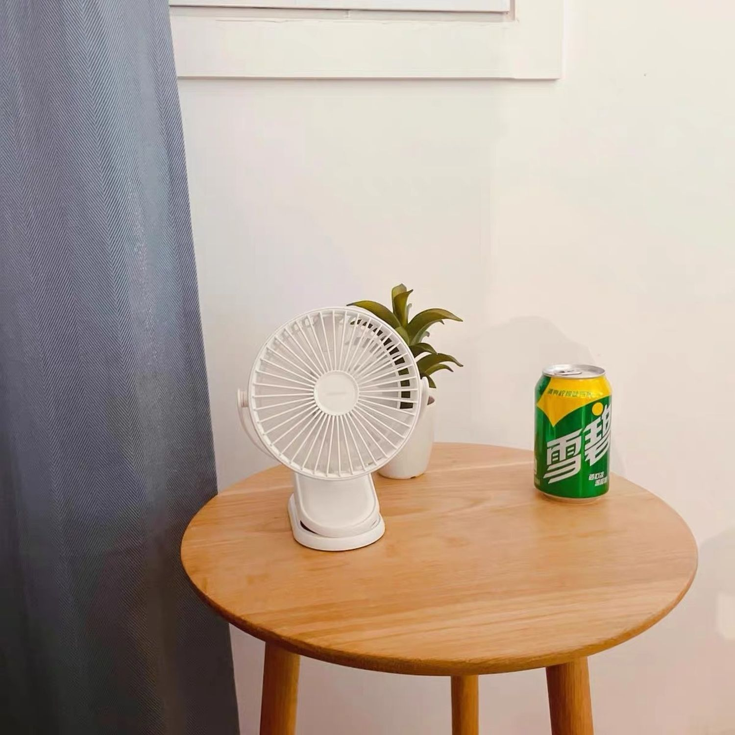 Ein weißer Ventilator auf einem Holztisch neben einer Limonadendose und einer kleinen Pflanze.