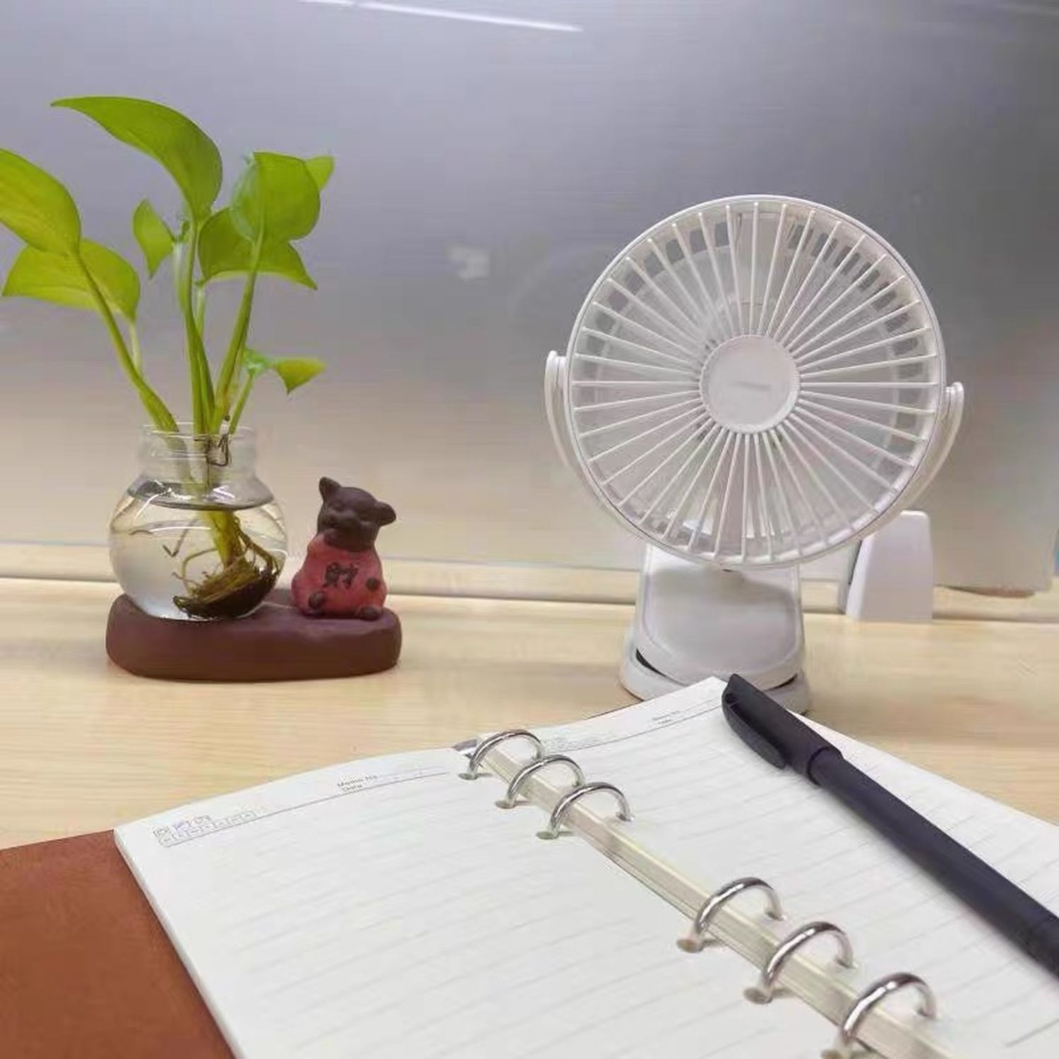 Schreibtisch mit Ventilator, Pflanze, Notizbuch und Stift. Hintergrund ist eine helle Wand.