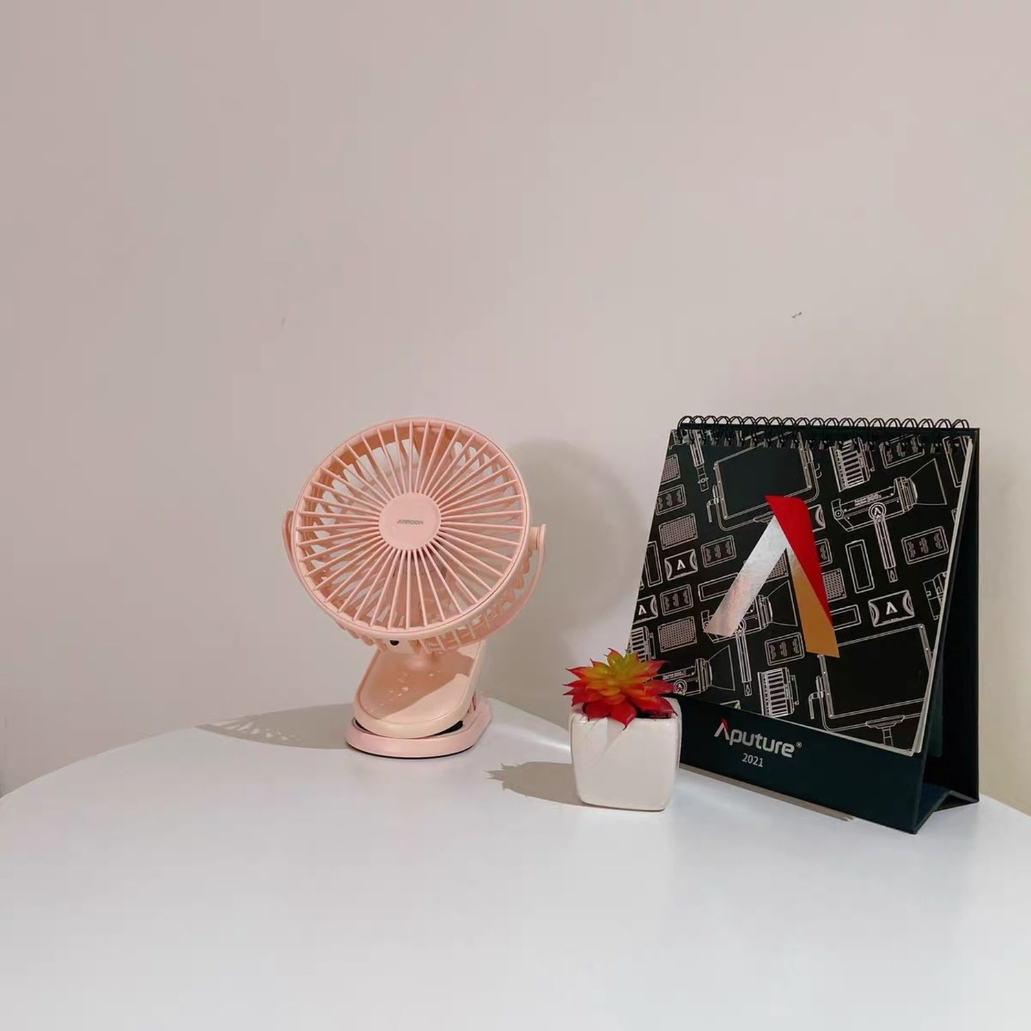 Rosa Ventilator, ein Kalender und eine kleine Topfpflanze auf einem weißen Tisch vor weißer Wand.