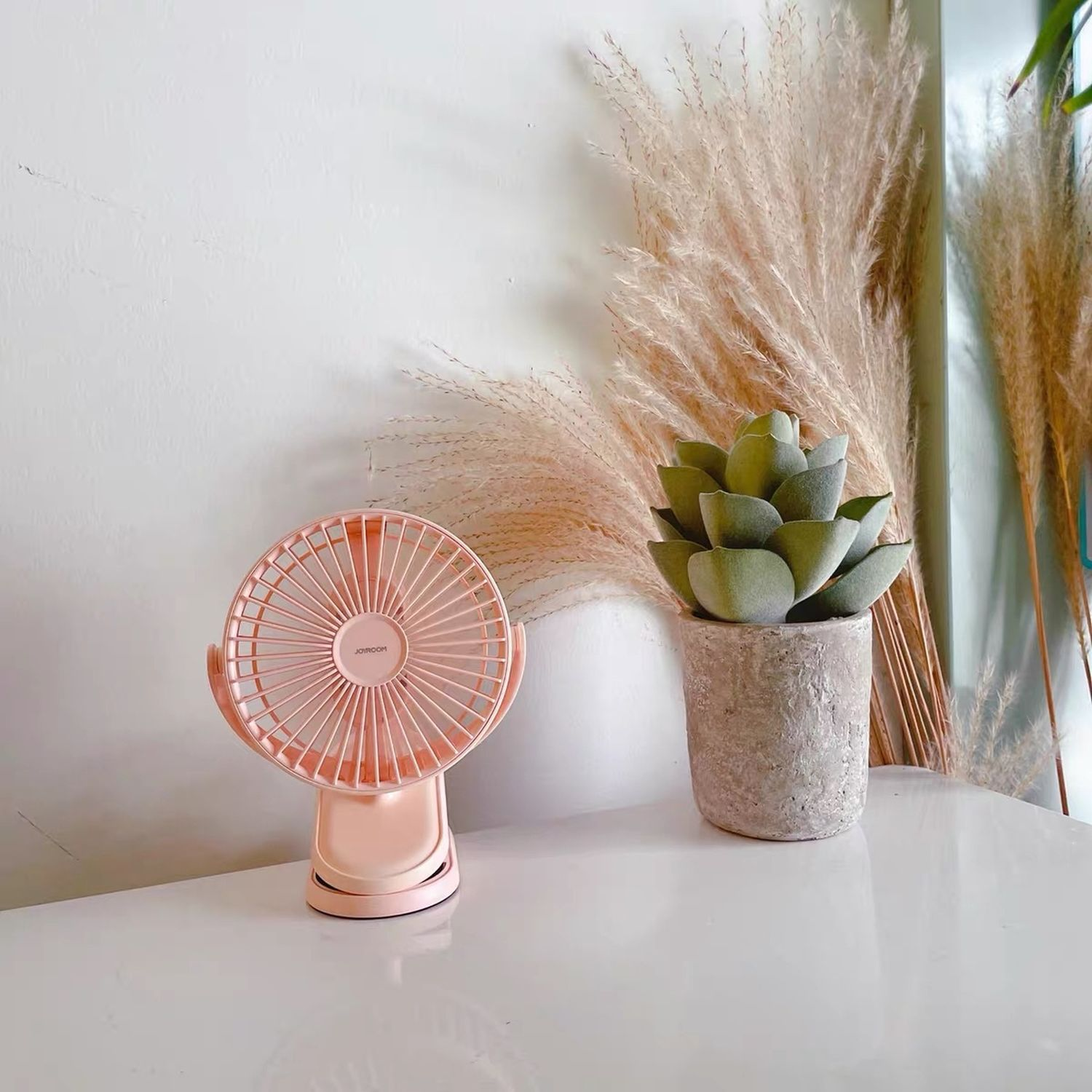 Rosa tragbarer Ventilator auf dem Tisch, mit einer Sukkulente und Pampasgras im Hintergrund.