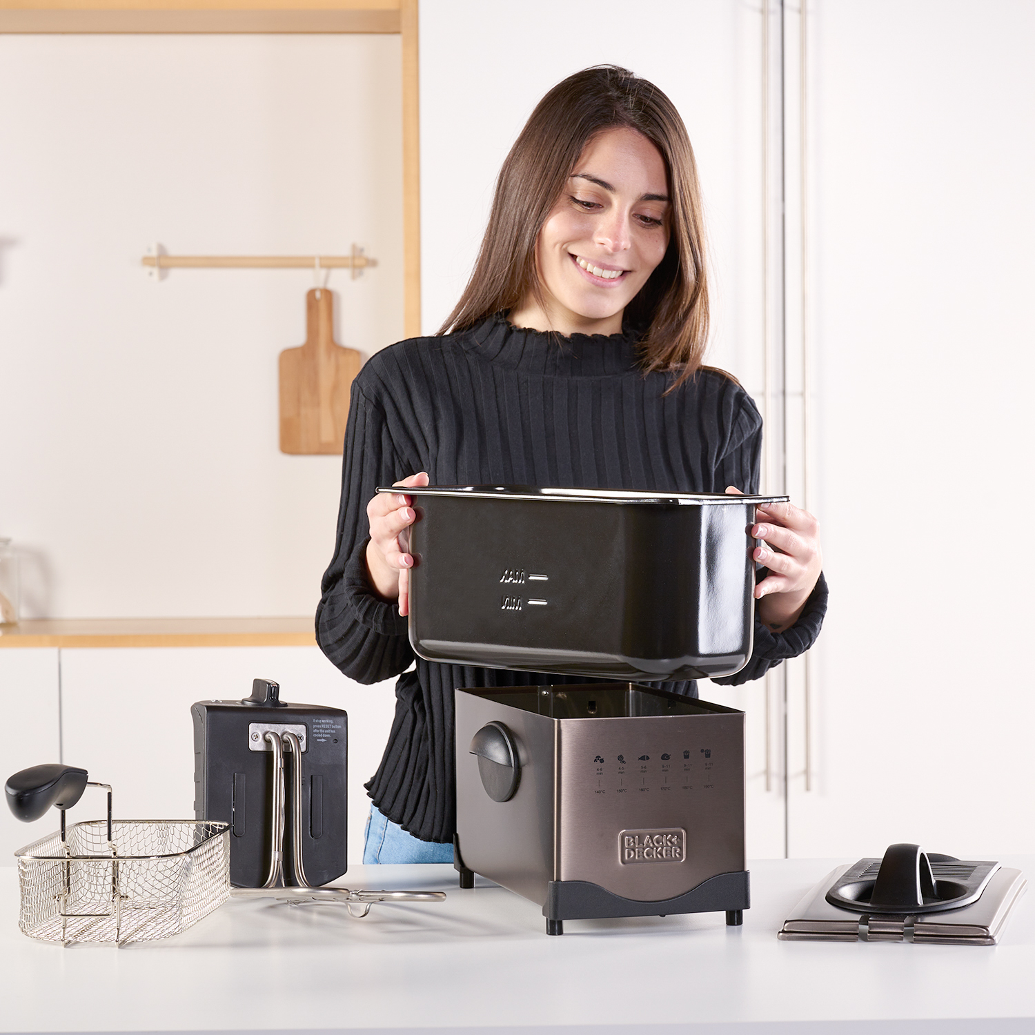 Kobieta trzyma czarną frytkownicę. Ma srebrny kosz i elementy sterujące. Tło jest białe.