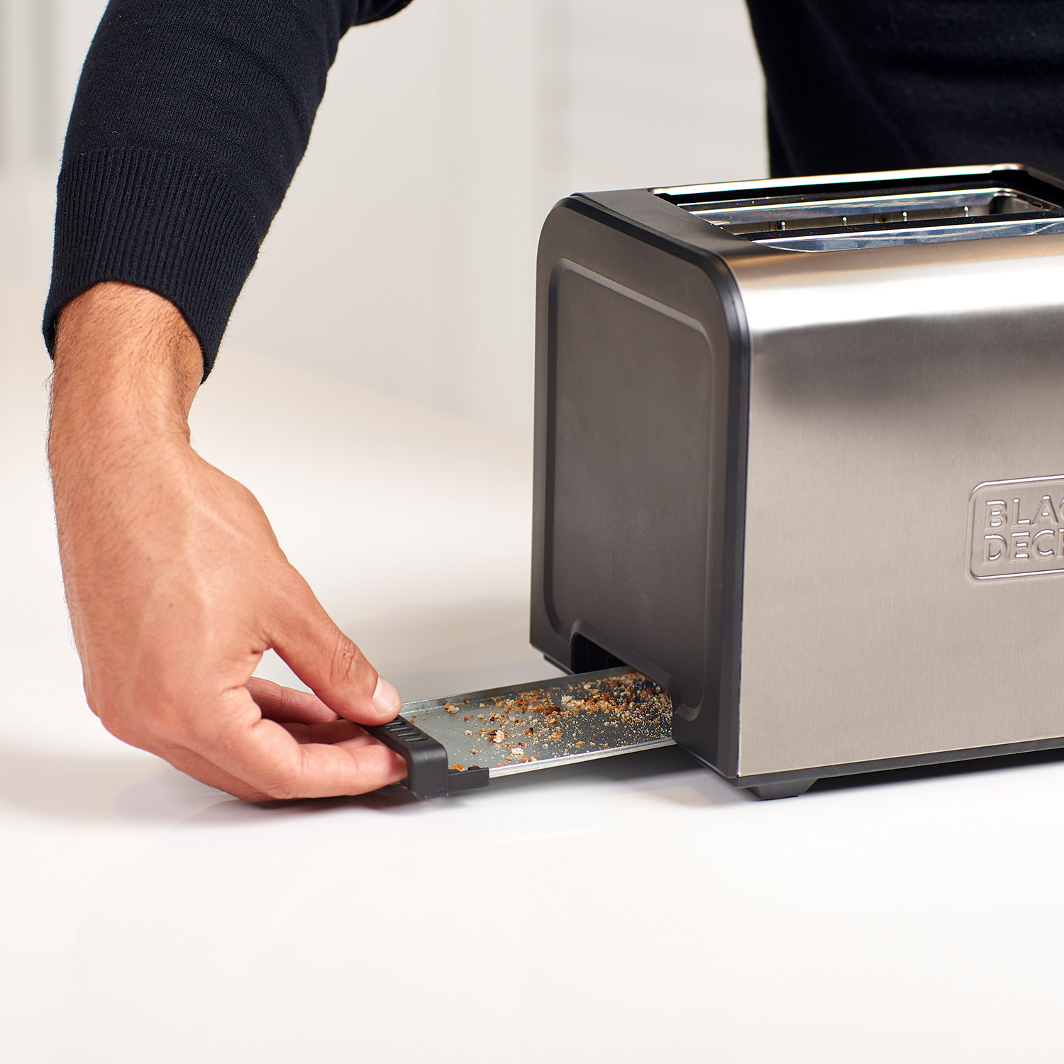 Osoba wyjmująca tackę na okruchy z srebrno-czarnego tostera na białej powierzchni.