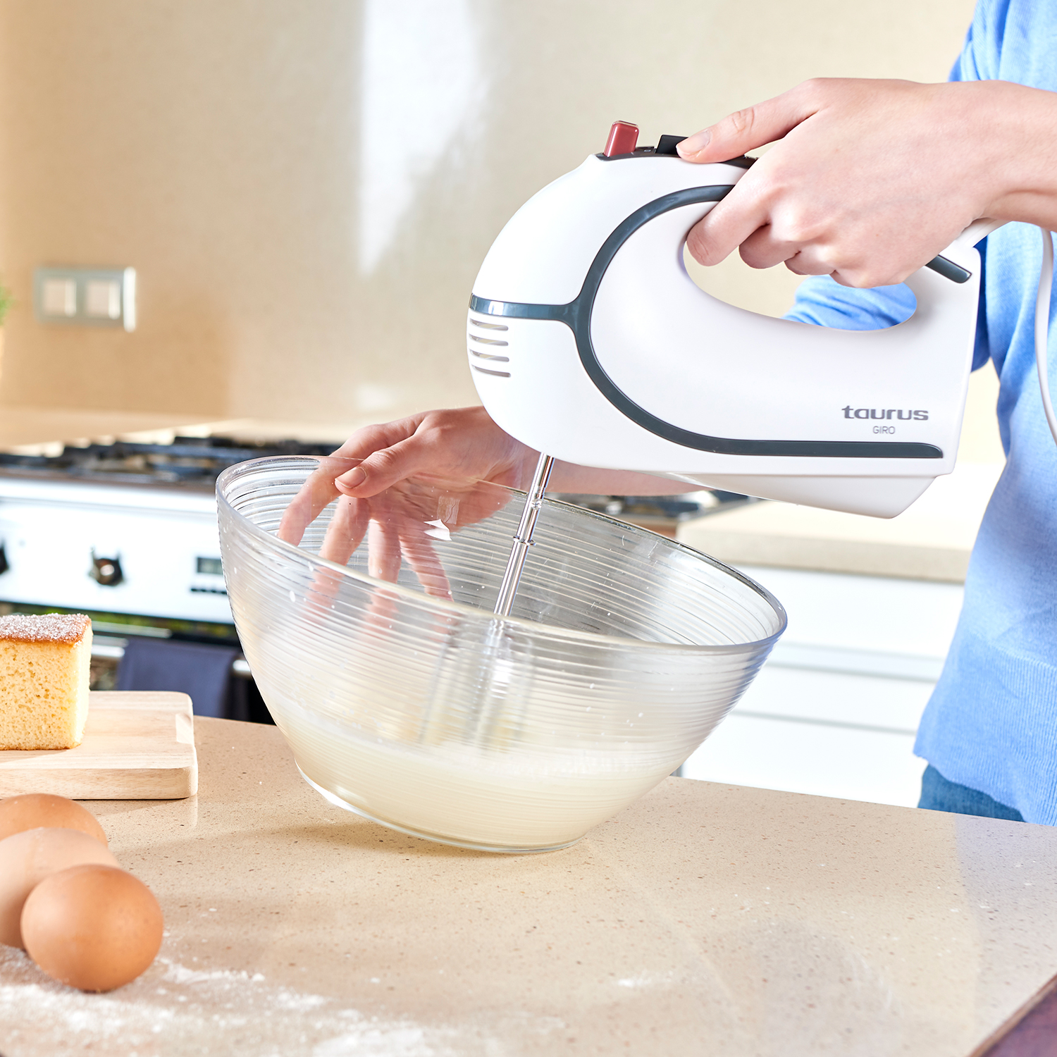 Una persona usa una batidora de mano blanca para batir ingredientes en un bol de vidrio sobre una encimera, con huevos cerca.