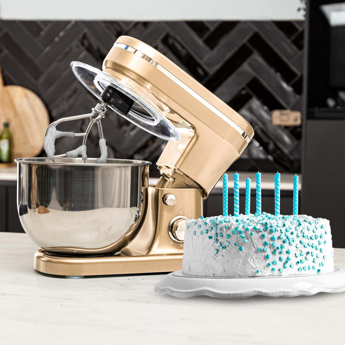 Gold stand mixer with a cake, candles, and a mixing bowl, on a black background.