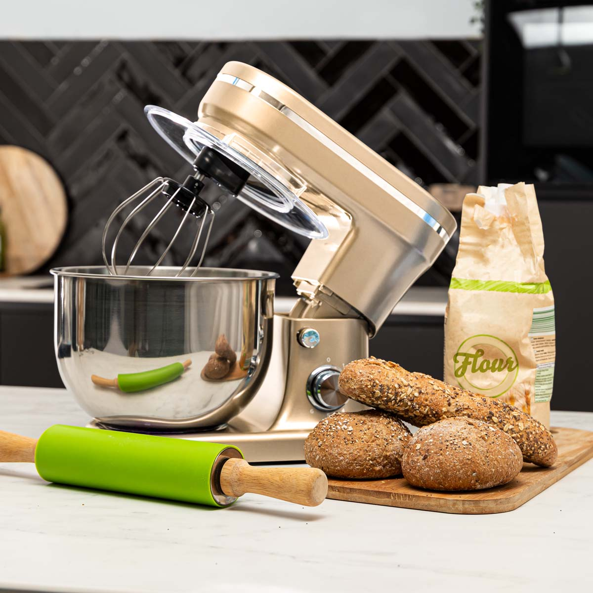 A gold KitchenAid mixer with baking supplies and bread rolls on a wooden board.