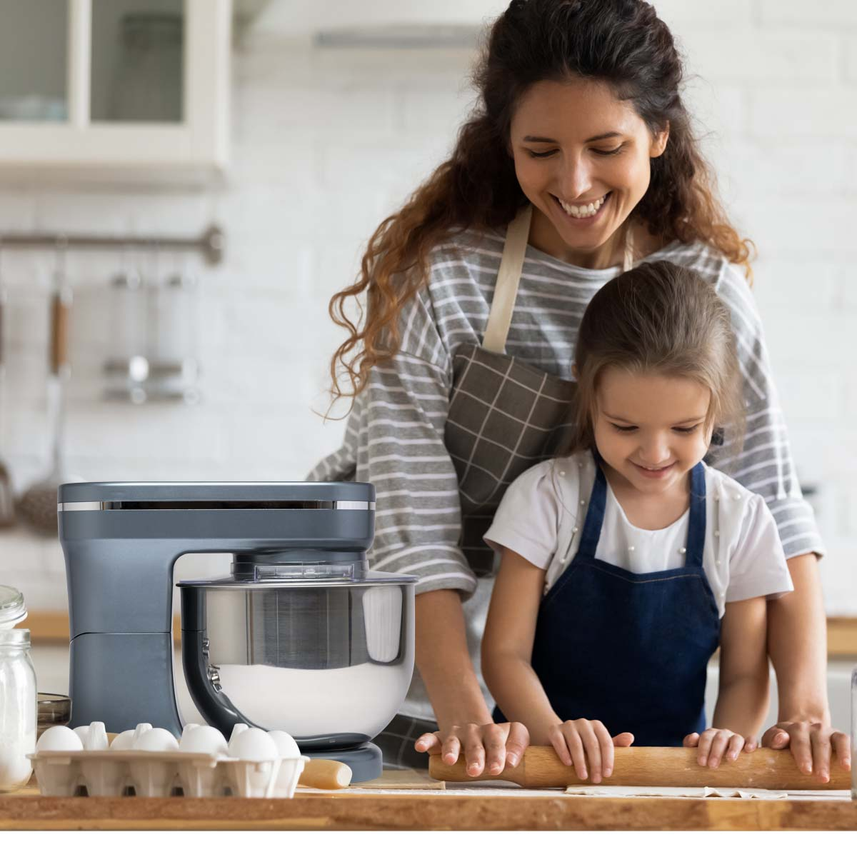 Woman and girl baking in a kitchen. There is a stand mixer, eggs, and a rolling pin on the counter.