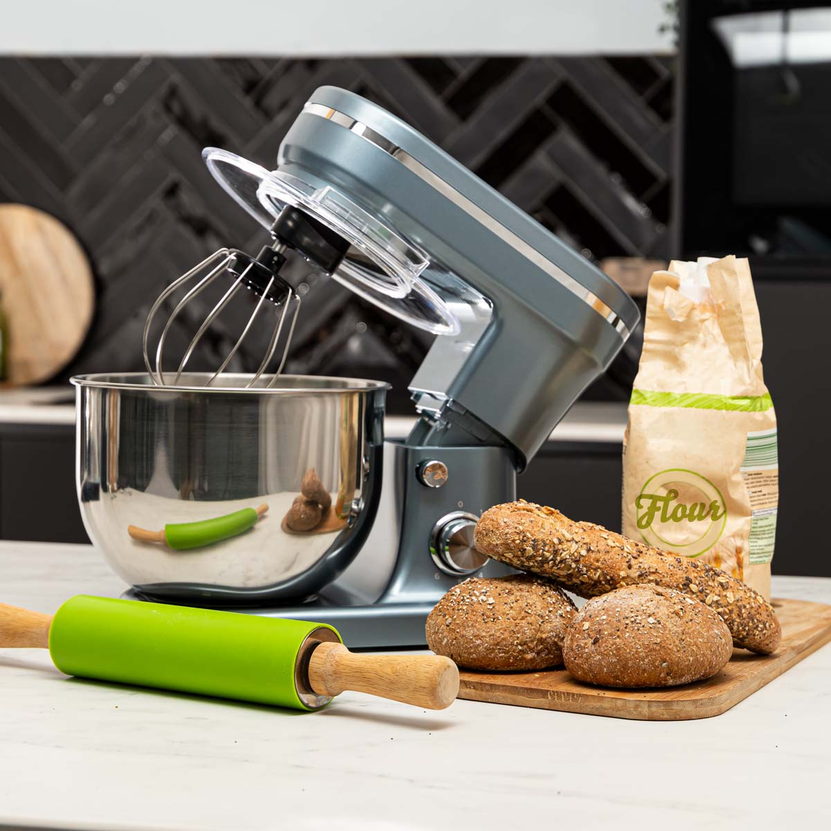 A stand mixer with a whisk attachment and a bowl, flour, rolling pin, and bread.