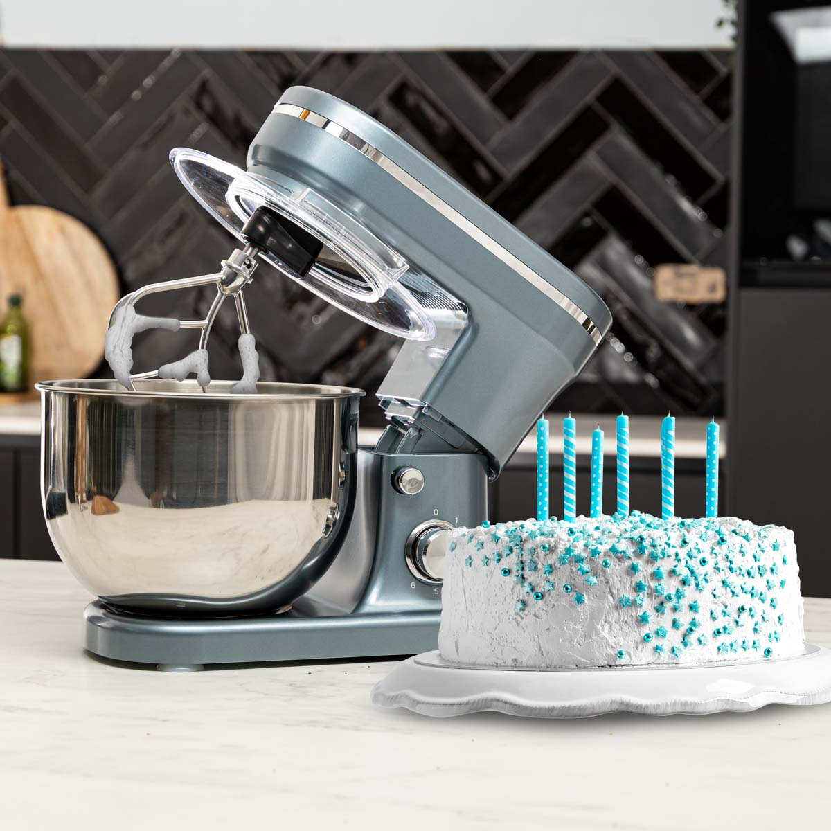 A grey stand mixer with a bowl and cake. Blue candles are on the cake, in front of a black wall.
