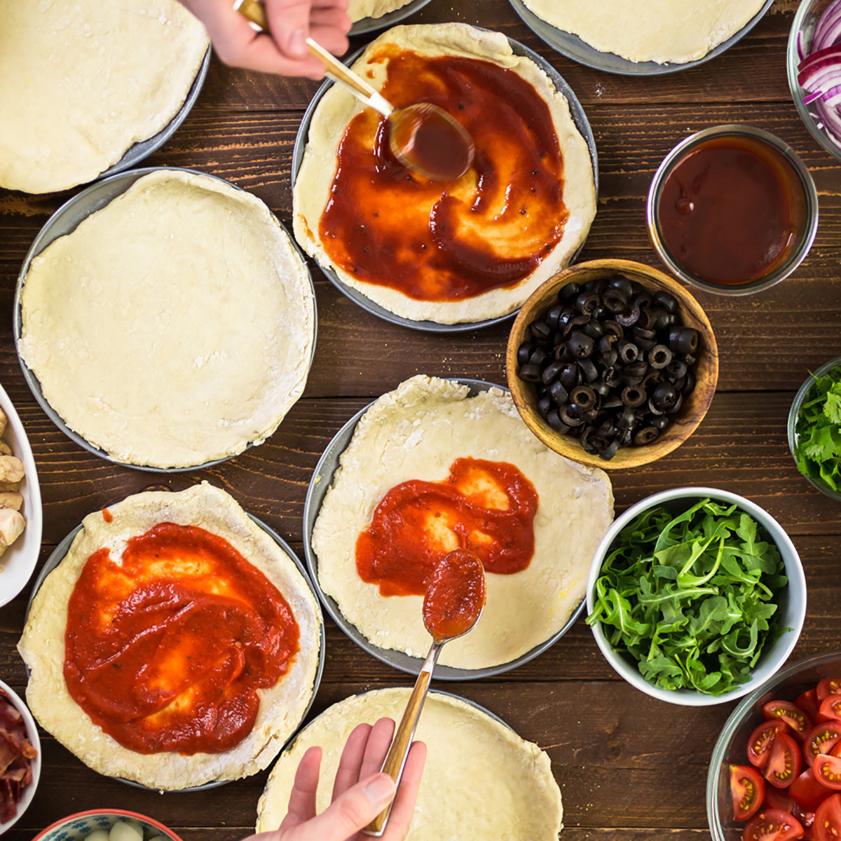 Handen die saus over pizzadeeg smeren. Ingrediënten: olijven, tomaten, rucola en uien.