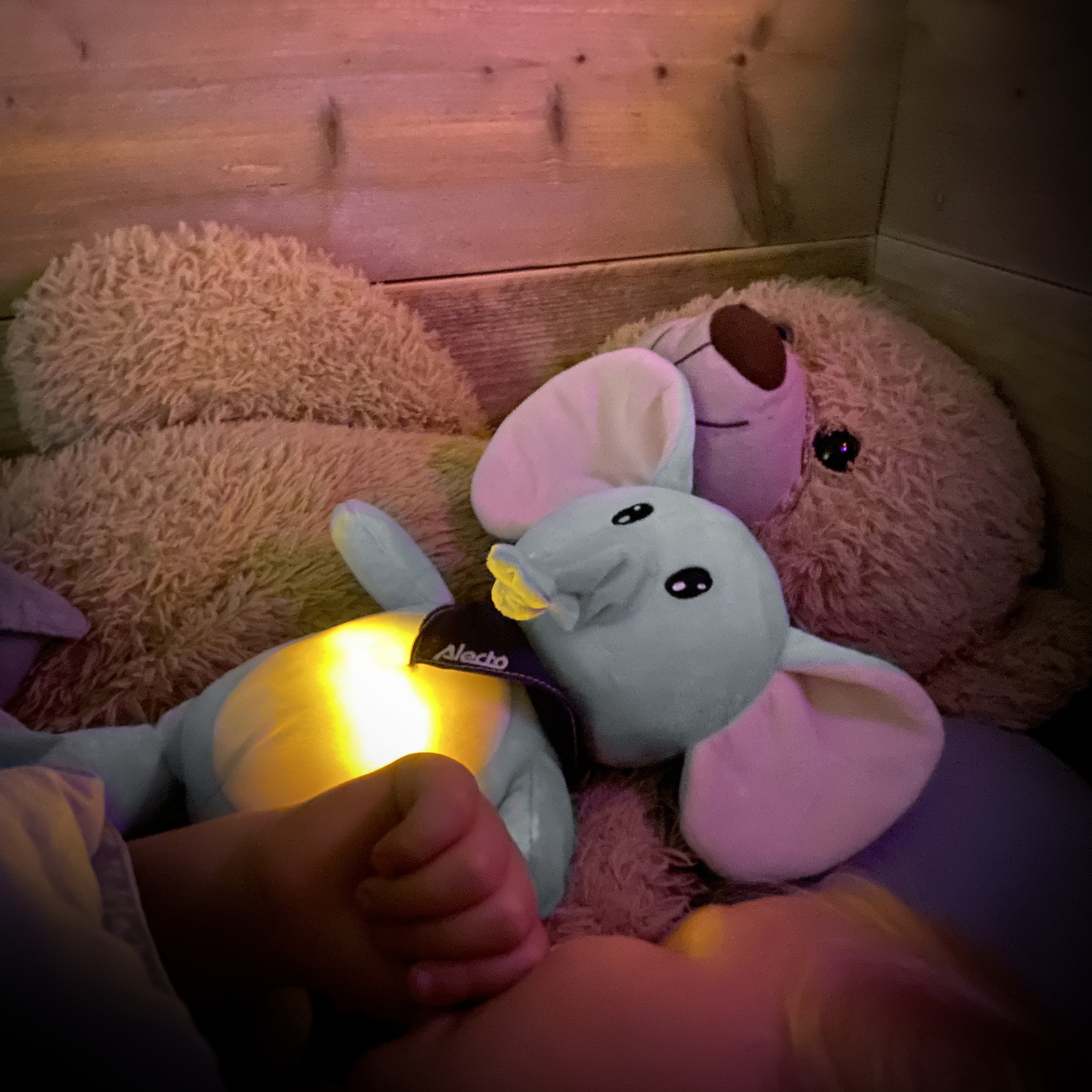Een kinderhand en een verlicht olifantenspeeltje naast een teddybeer in een houten bed.