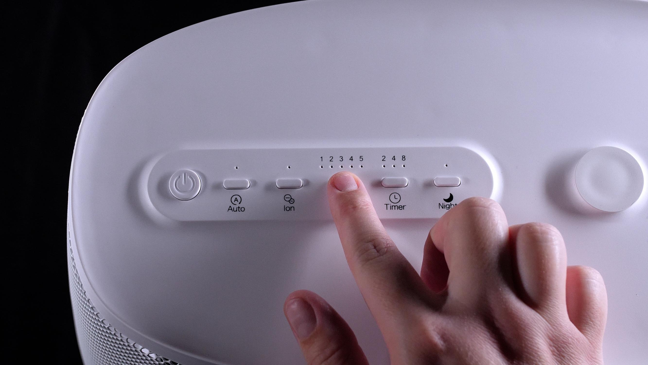 Una mano preme un pulsante su un purificatore d'aria bianco. I pulsanti includono Auto, Ion, Timer e Notte.