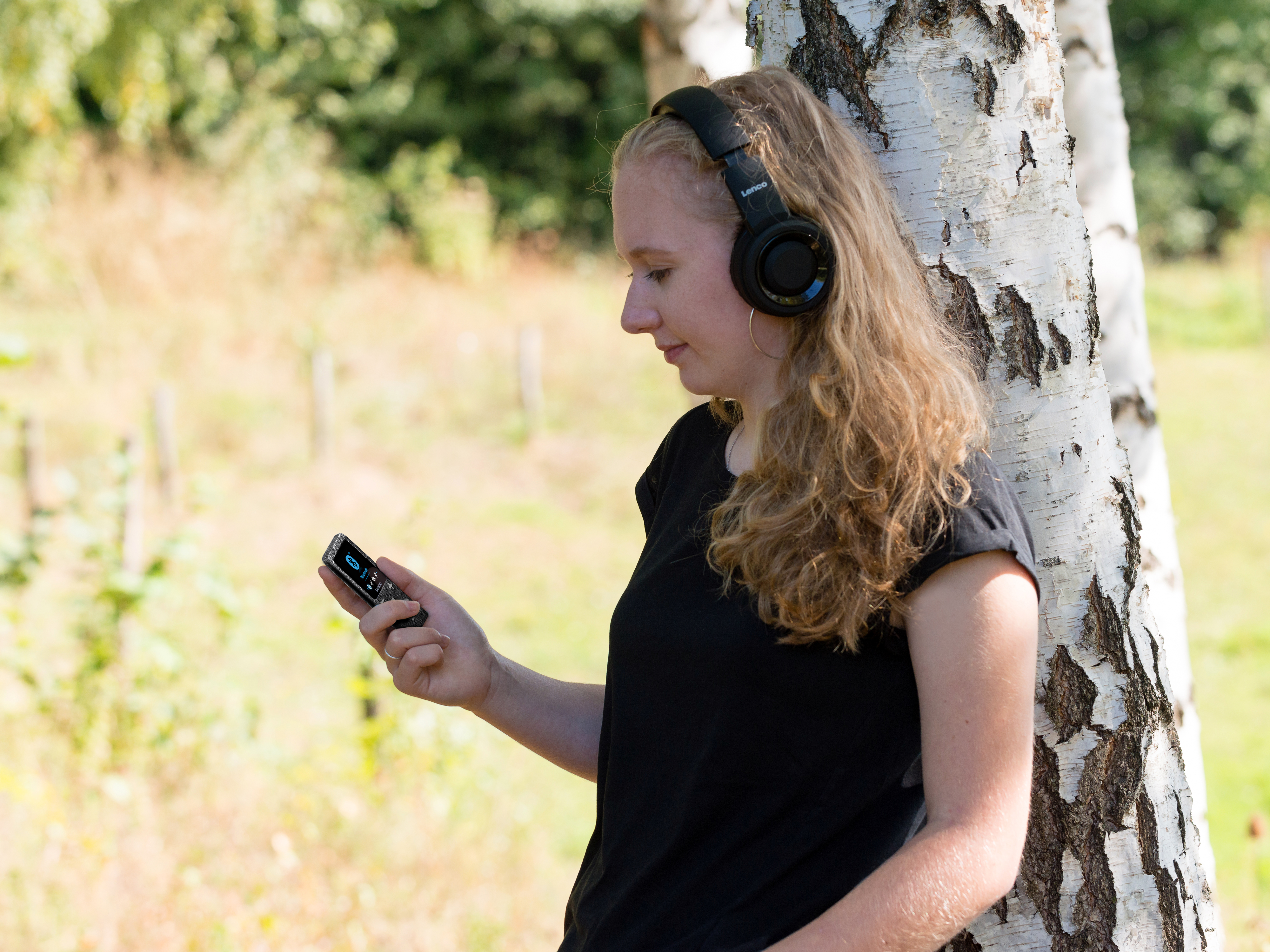 Kobieta w słuchawkach, trzymająca telefon, stojąca w pobliżu drzewa w naturze.