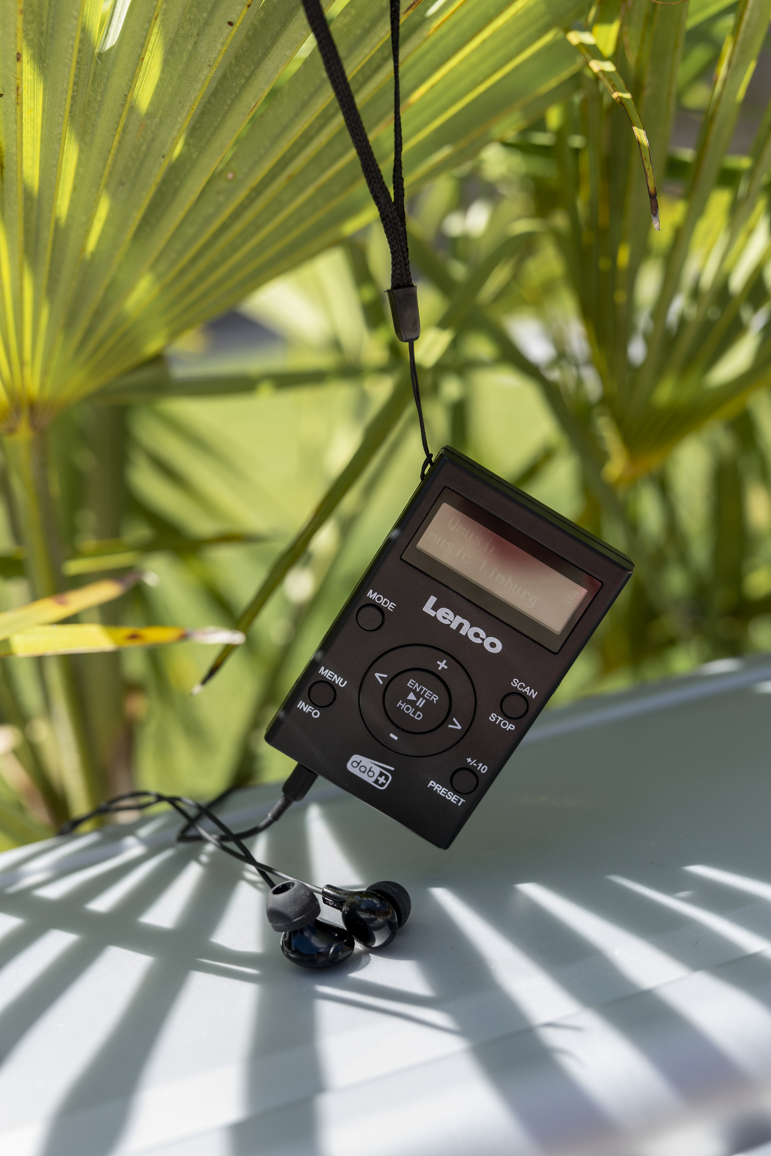 Ein schwarzes digitales Lenco-Radio mit Bildschirm. Das Radio hängt mit Kopfhörern vor grünen Blättern.