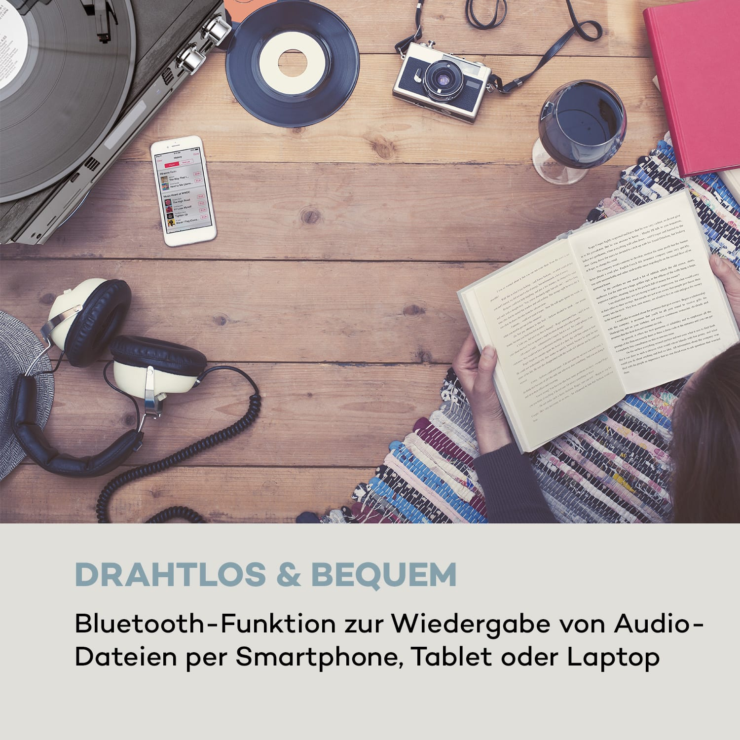 Person liest Buch, mit Kopfhörern, Plattenspieler und Smartphone auf Holztisch.