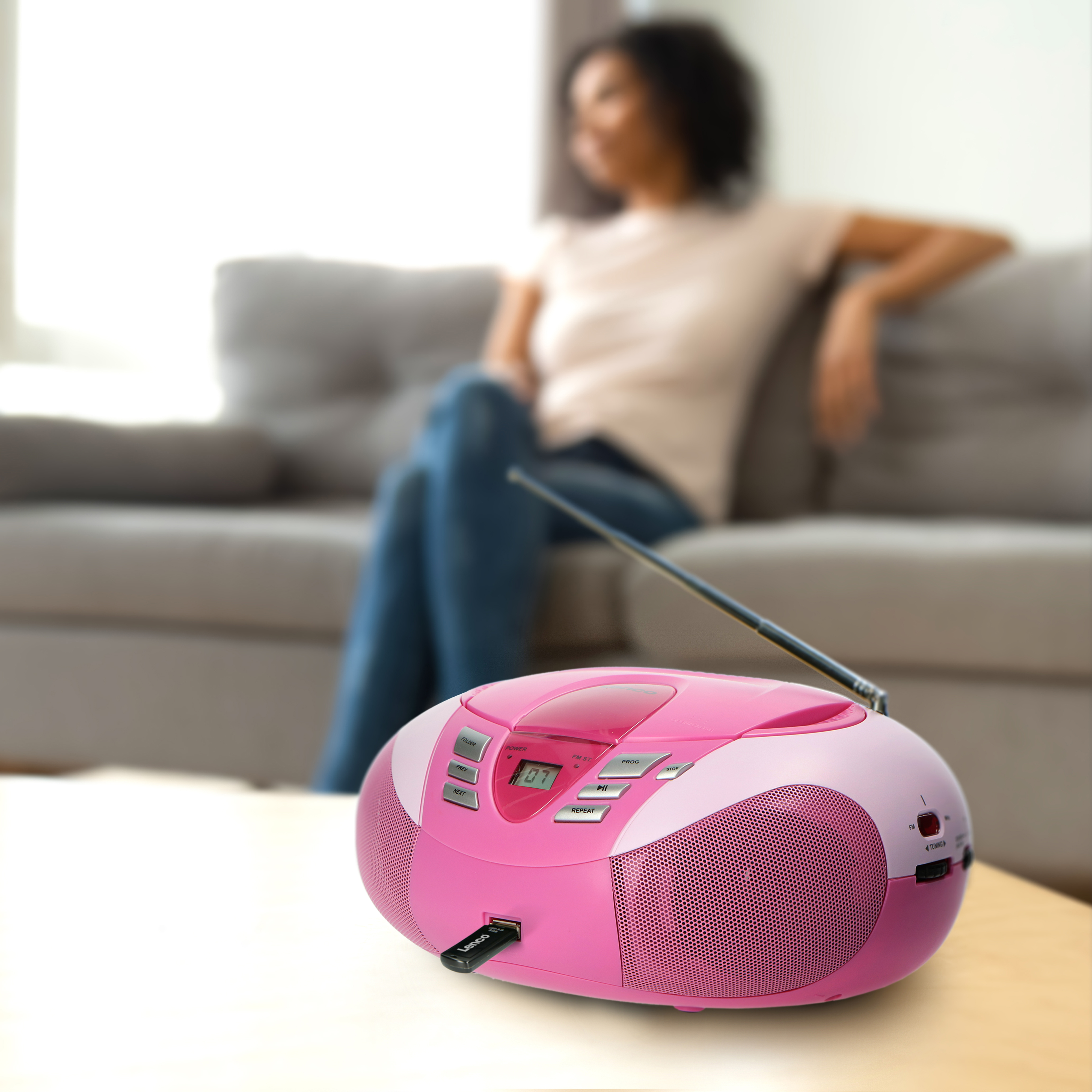 Ein pinkfarbenes Boombox-Radio mit eingestecktem USB-Stick auf einem Tisch, Frau im Hintergrund.