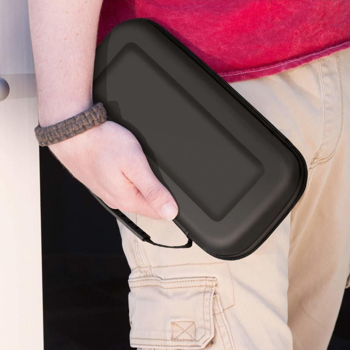 Eine Person hält einen schwarzen, rechteckigen Koffer. Sie trägt khakifarbene Shorts und ein rotes Hemd.