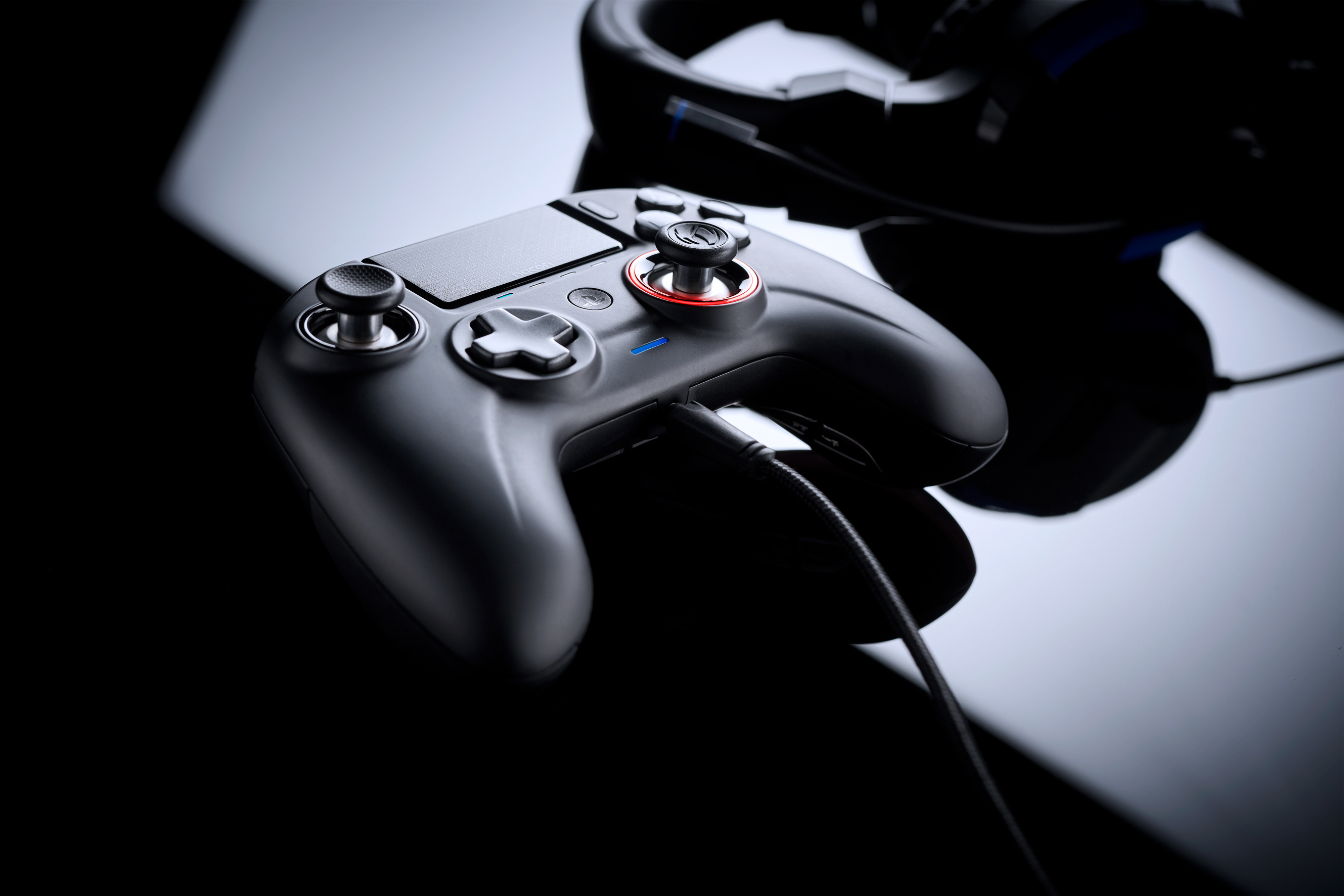 Mando de juegos con cuerpo negro, cable negro y auriculares en el fondo, todo sobre superficie reflectante.