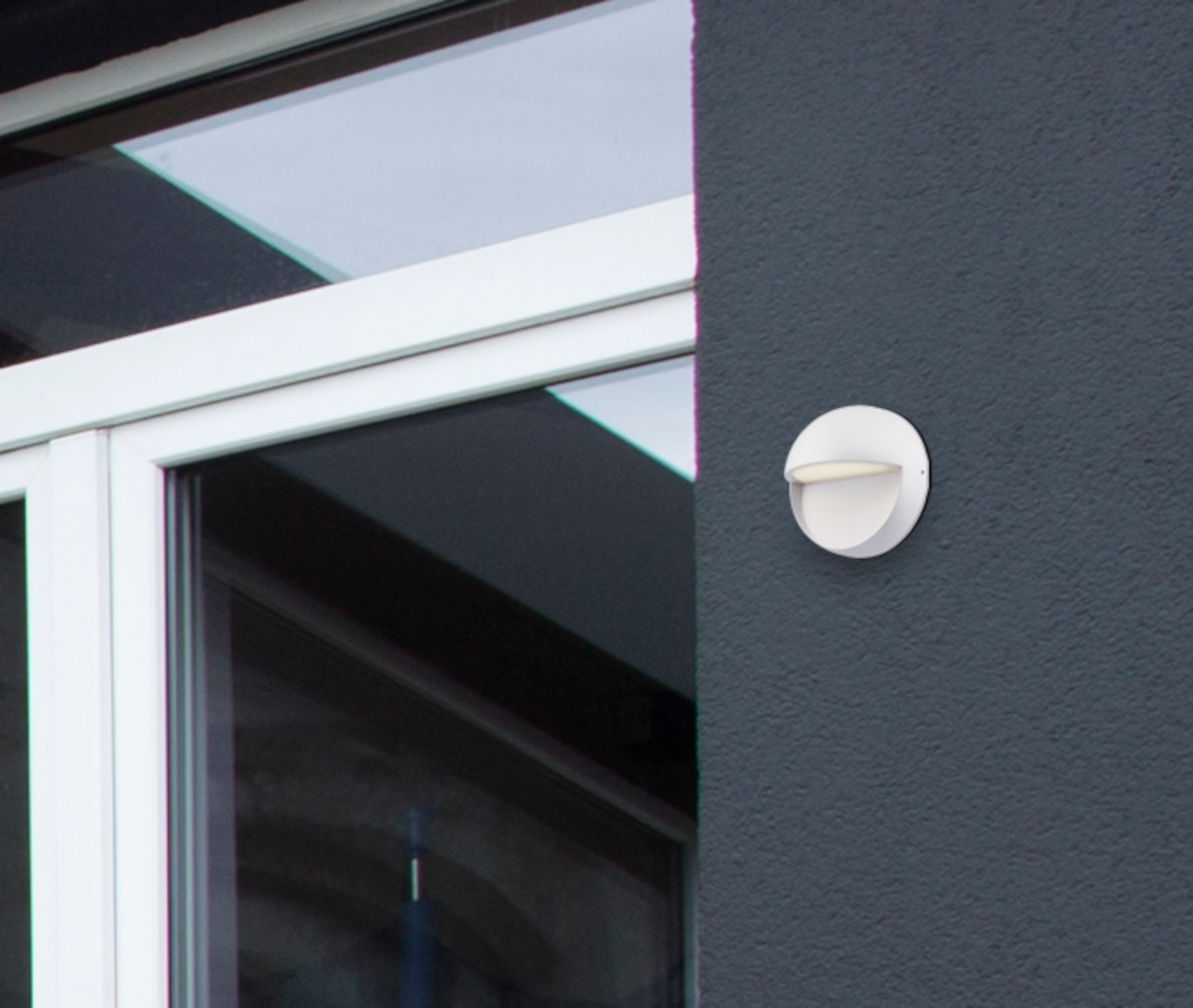 Weiße, kreisförmige Außenleuchte an einer dunkelgrauen Wand in der Nähe eines Fensterrahmens montiert.