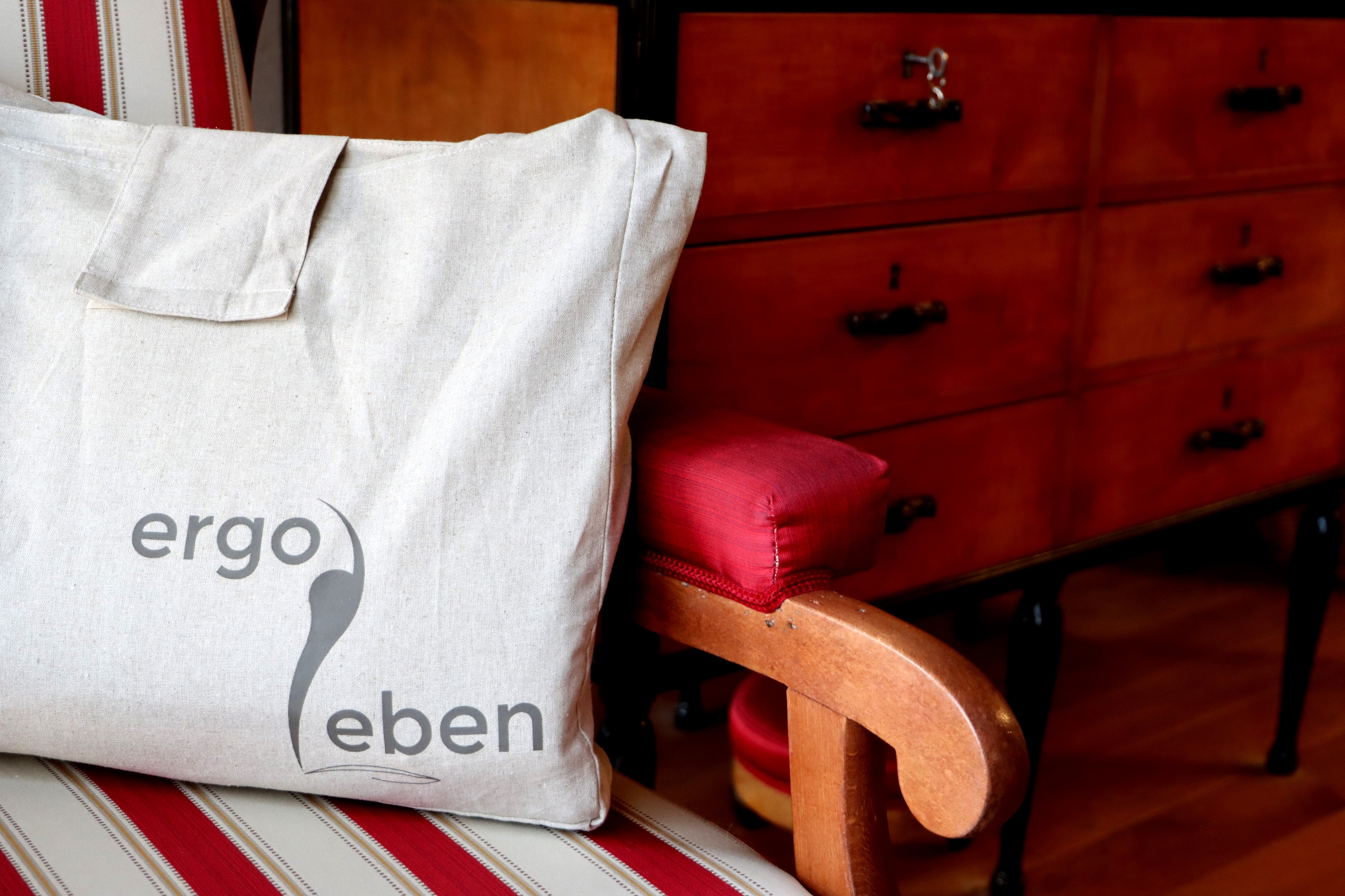 Eine cremefarbene Tragetasche mit den Worten „ergo leben“ und einem grauen Logo auf einem gestreiften Stuhl.