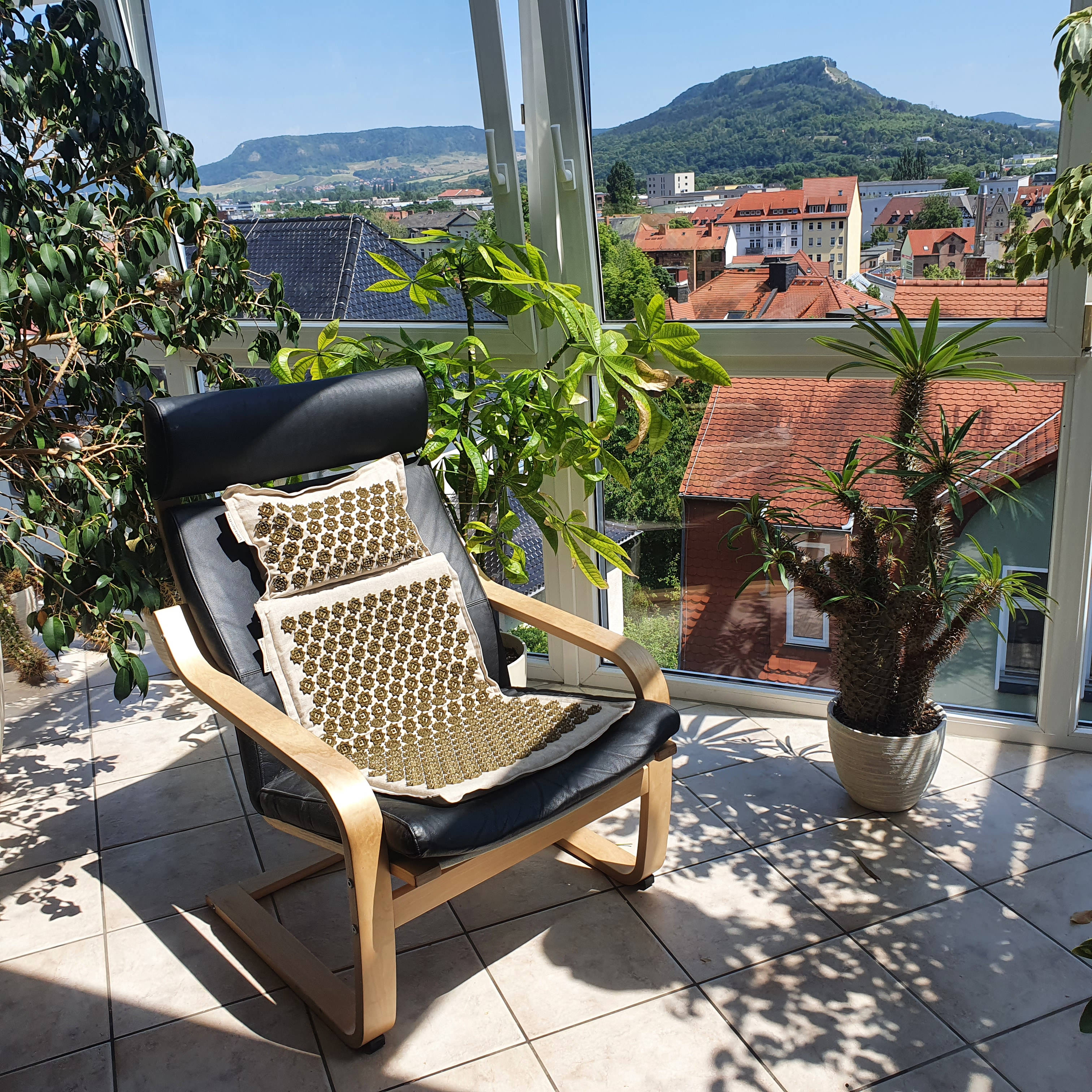 Wintergarten mit Stuhl, Kissen und Pflanzen. Blick auf Gebäude und Berge aus den Fenstern.