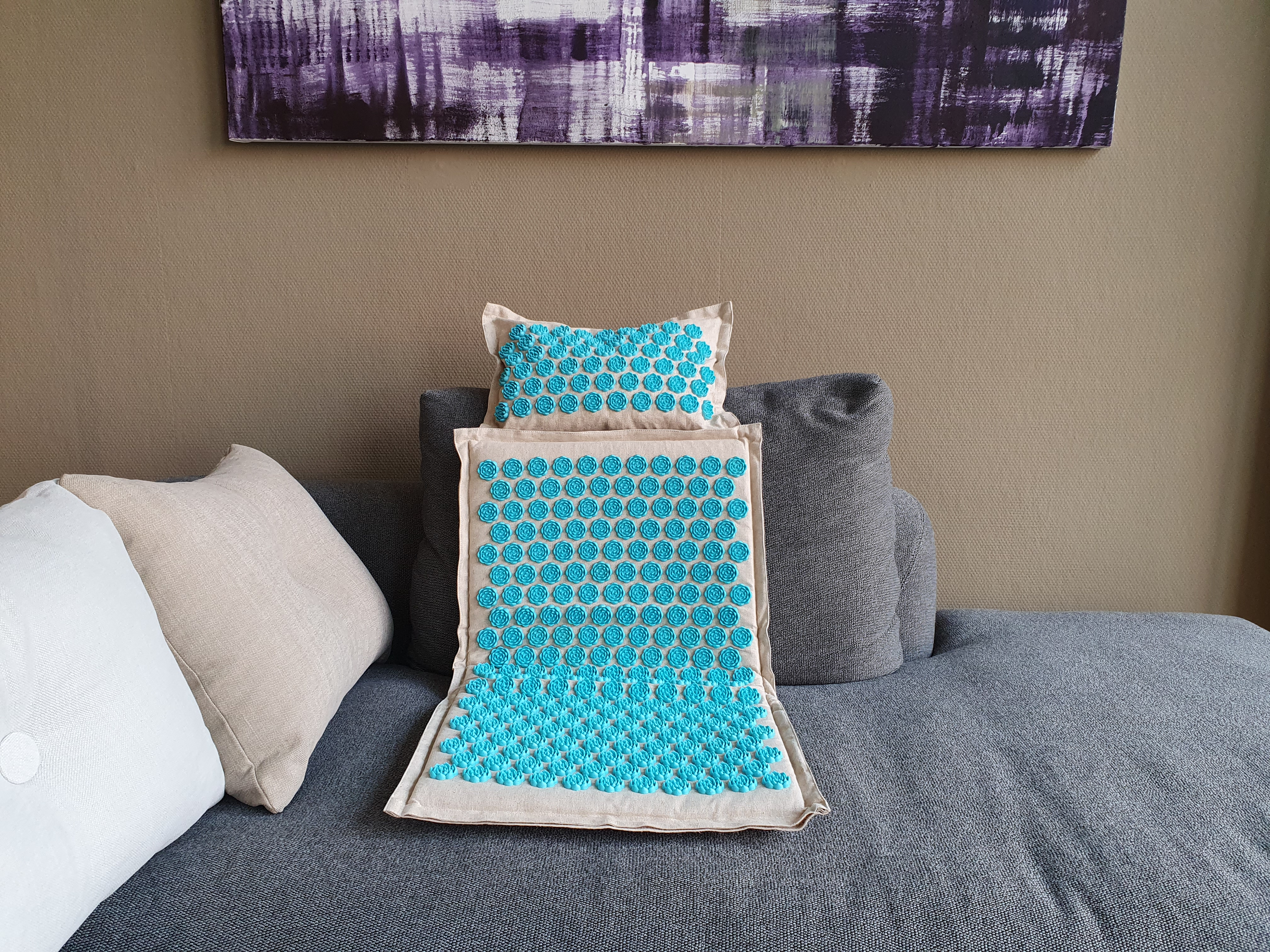 Blaue, stachelige Massagematte und Kissen auf einem grauen Sofa mit weißen und grauen Kissen.