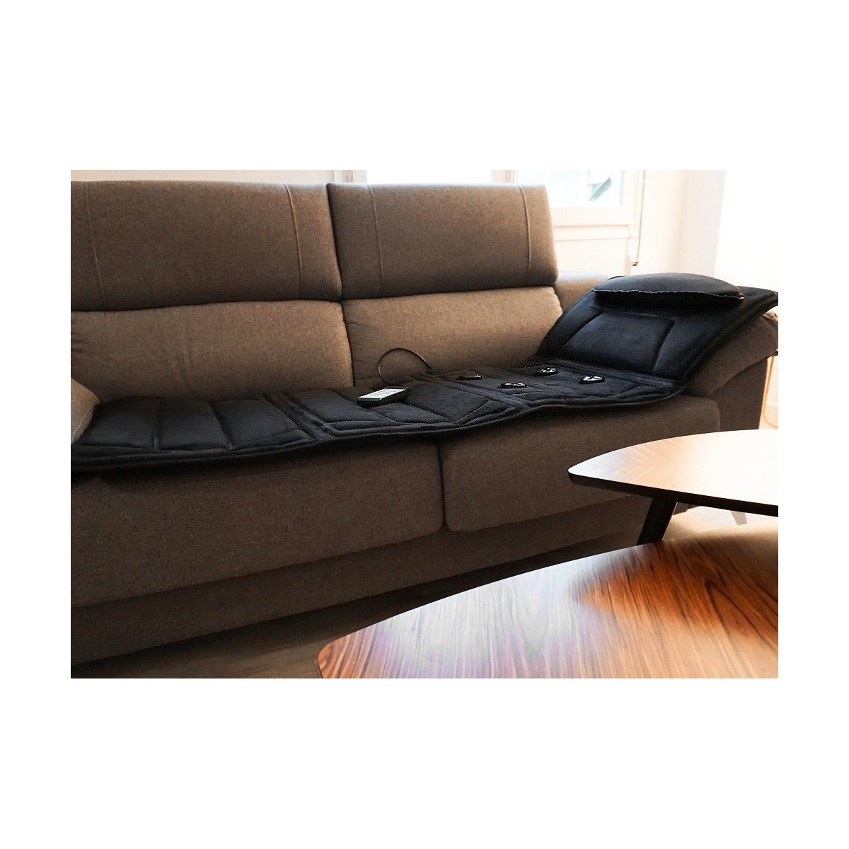 Schwarze Massagematte auf einem braunen Sofa. Rechts ein Holztisch.