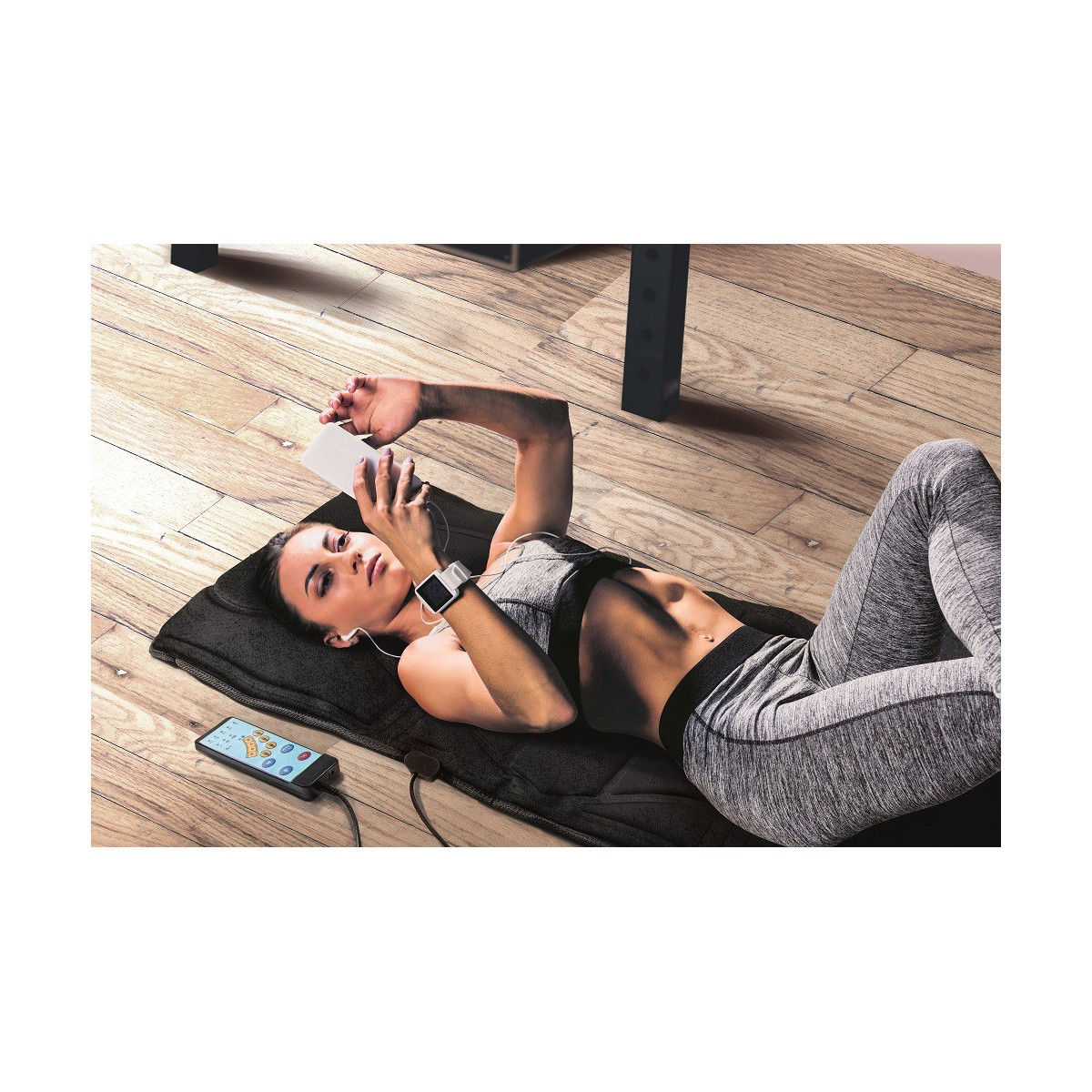 Frau liegt auf einer Matte, benutzt Telefon und Smartwatch. Sie trägt Trainingskleidung.