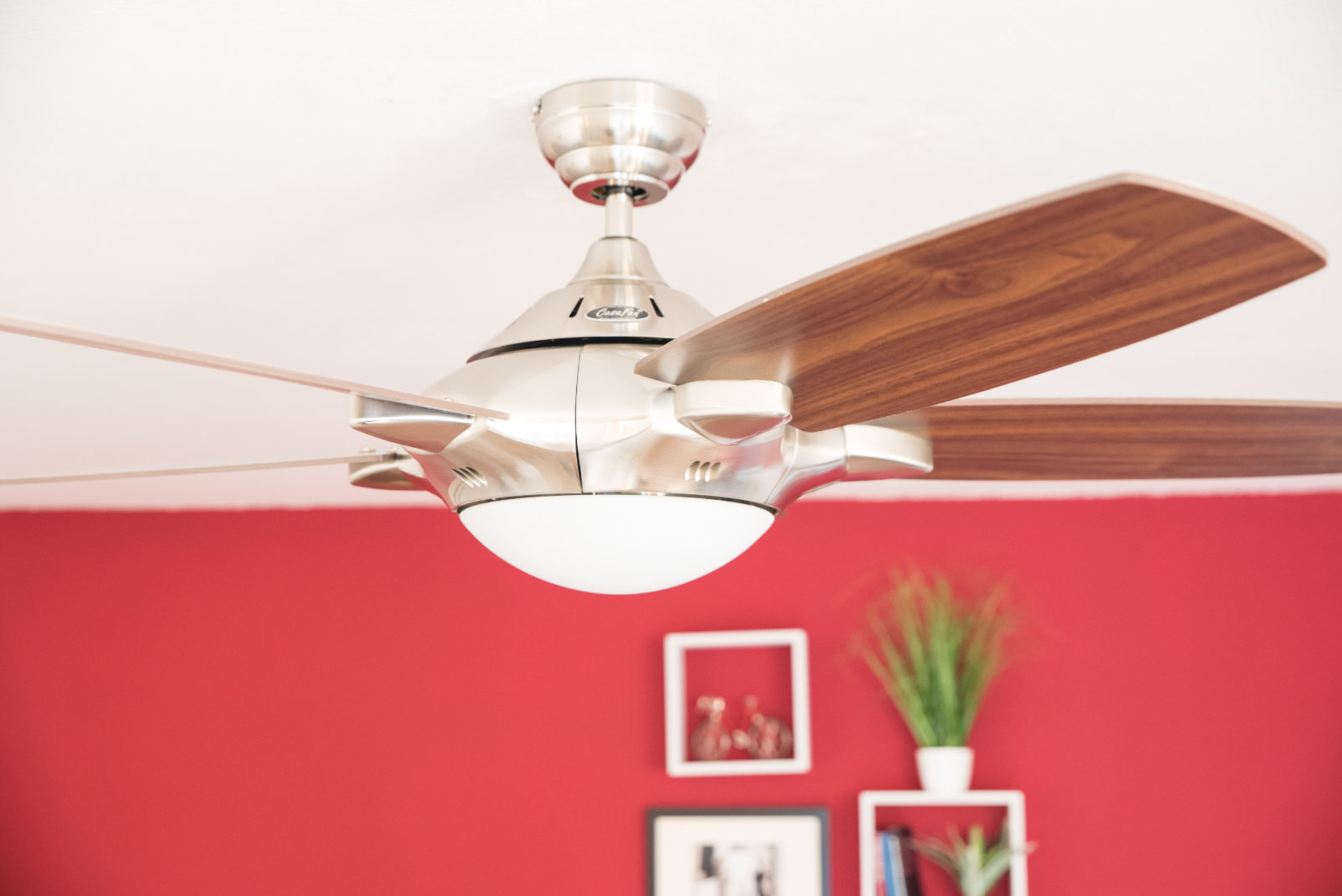 Ein Deckenventilator mit braunen Flügeln, an einer weißen Decke vor einer roten Wand montiert.