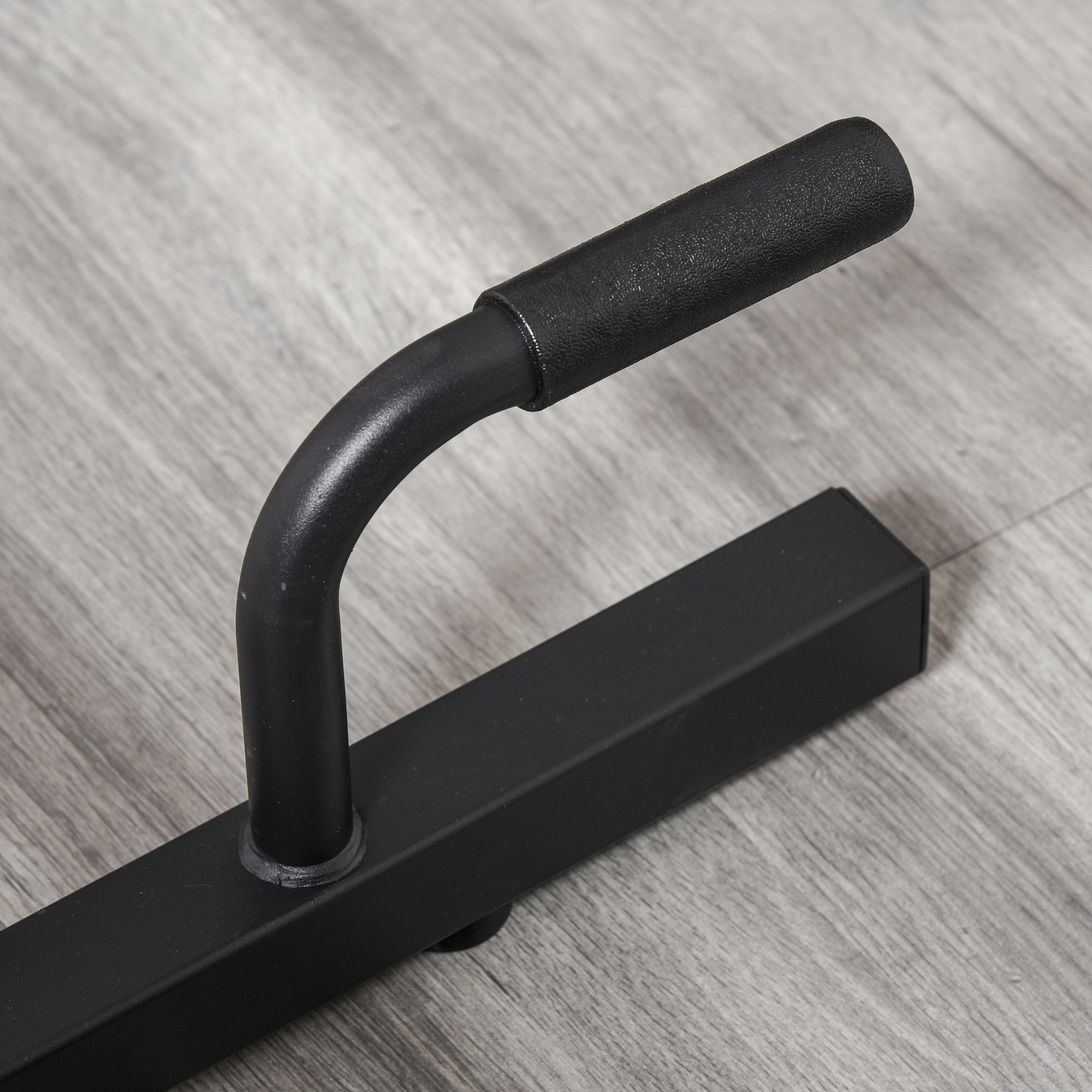 Barra per flessioni nera su un pavimento in legno. Ha una maniglia curva con impugnatura nera.