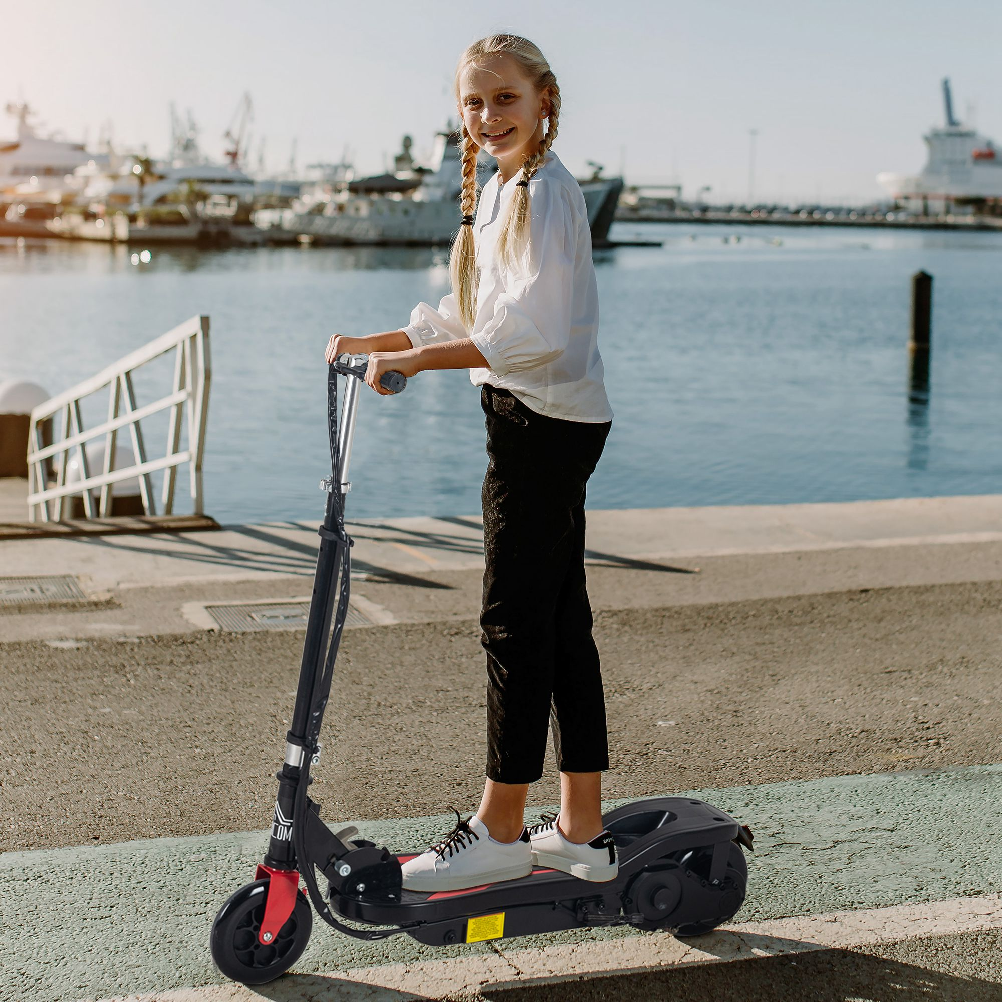 Niña montando un patinete eléctrico negro y rojo en un paseo marítimo pavimentado.