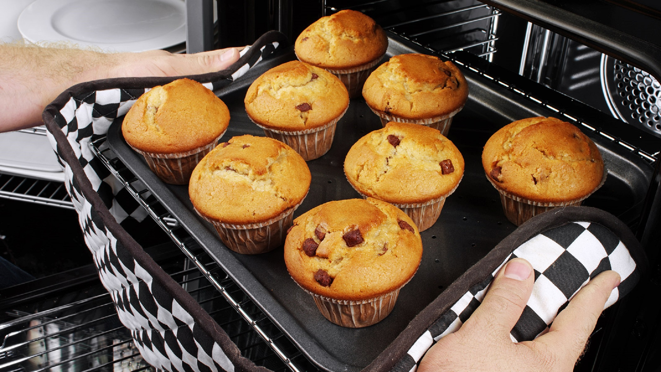 Manos con guantes de horno sacando una bandeja de muffins horneados. Los muffins son dorados con chispas de chocolate.
