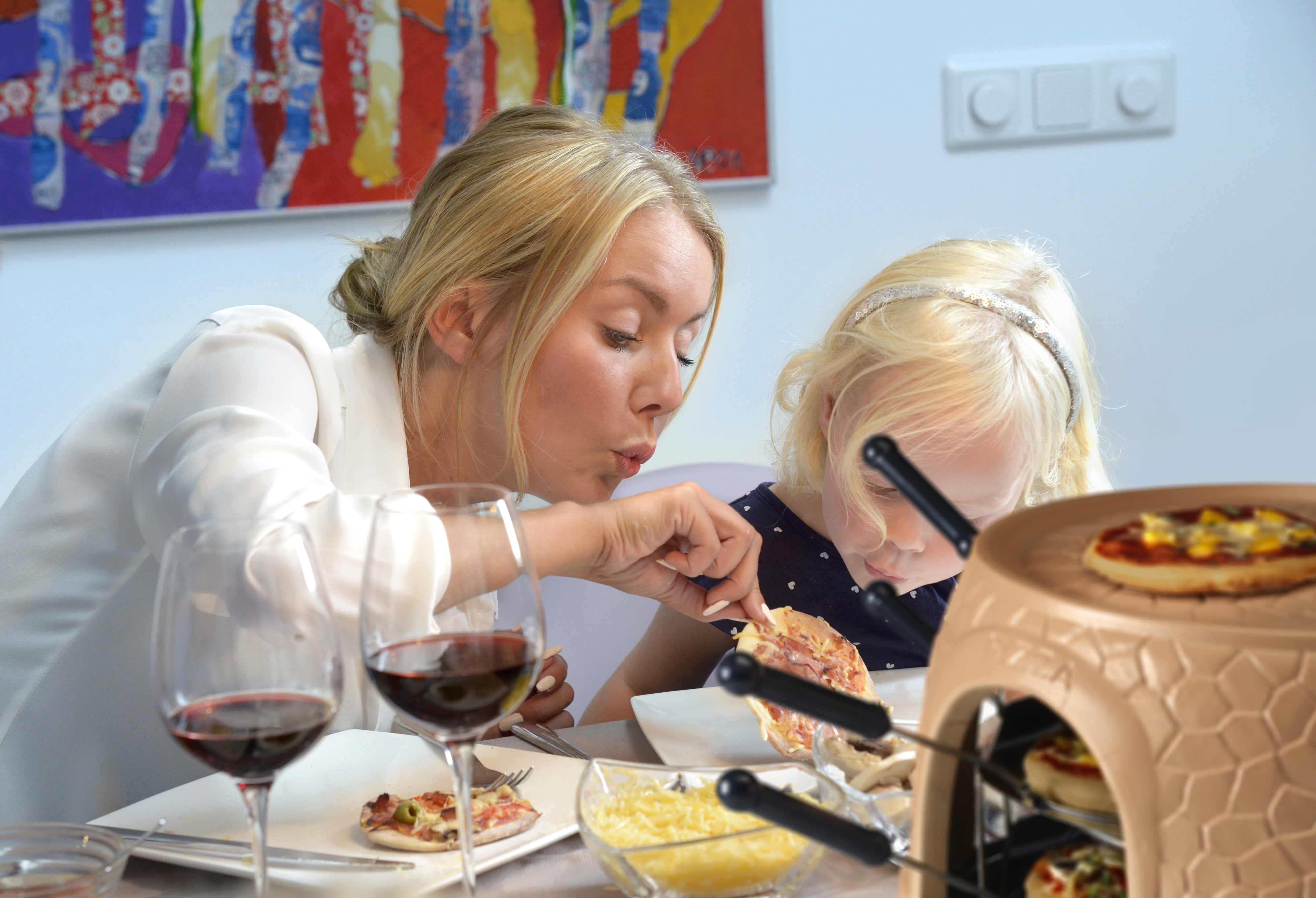 Donna e bambino mangiano pizza da un piccolo forno per pizza. Sono presenti calici e formaggio.