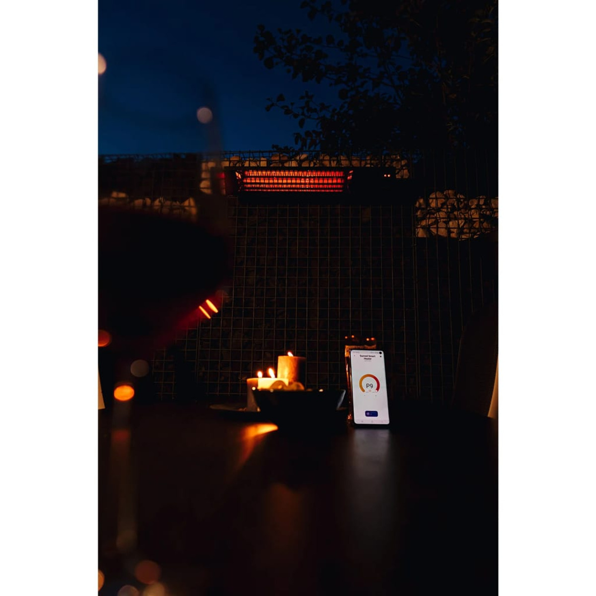 Un radiateur extérieur allumé avec un téléphone affichant les paramètres, des bougies et un bol de nourriture sur une table la nuit.
