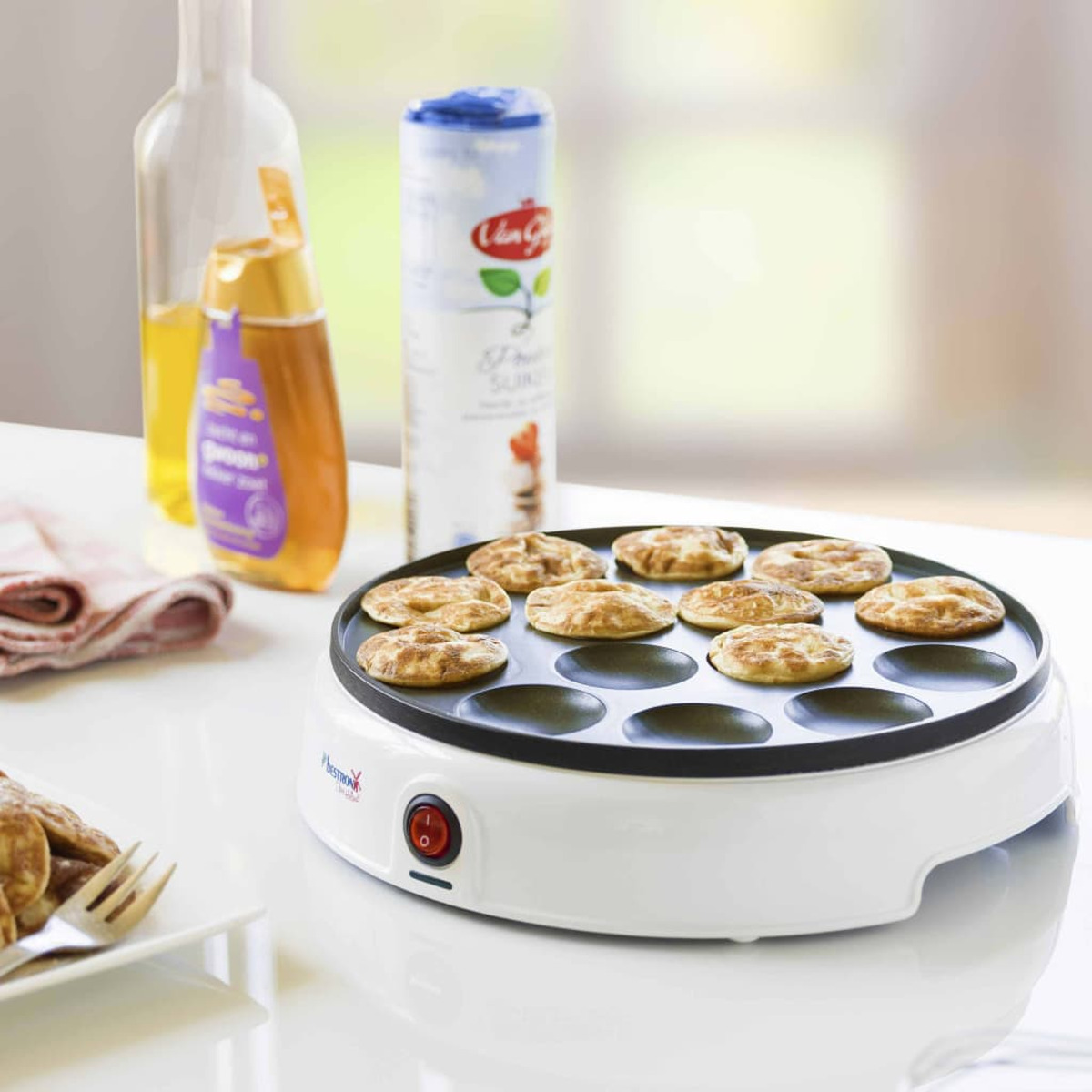 Białe urządzenie do robienia naleśników z gotowymi naleśnikami, butelkami i składnikami na białej powierzchni.